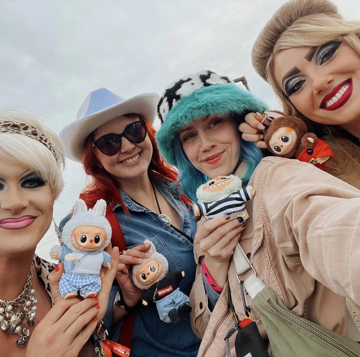 Four women smiling outdoors, holding plush toys and wearing colorful hats and makeup.