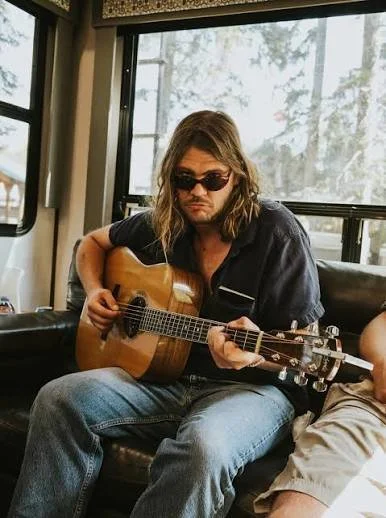 A man with long hair and sunglasses playing an acoustic guitar while sitting on a black leather couch indoors near a window.