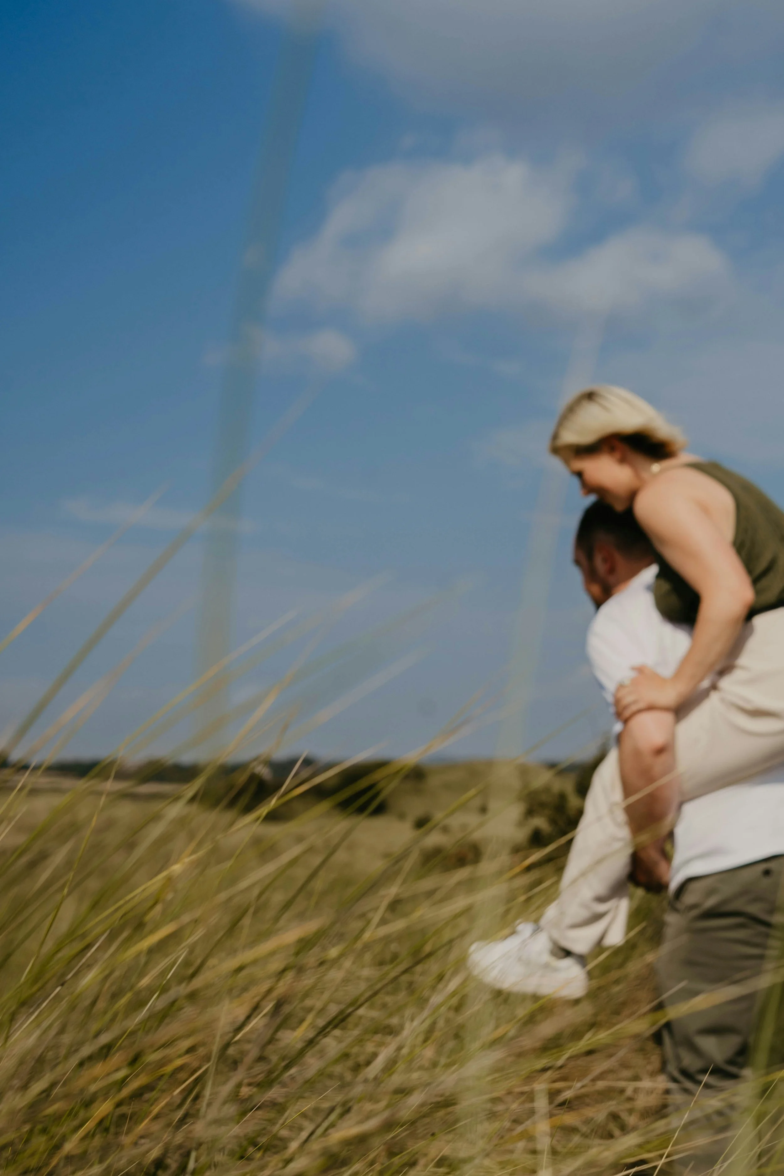 Woman on partner’s back, walking through tall grass under a clear sky — symbolizing safety, trust, and lightheartedness in holistic partnership