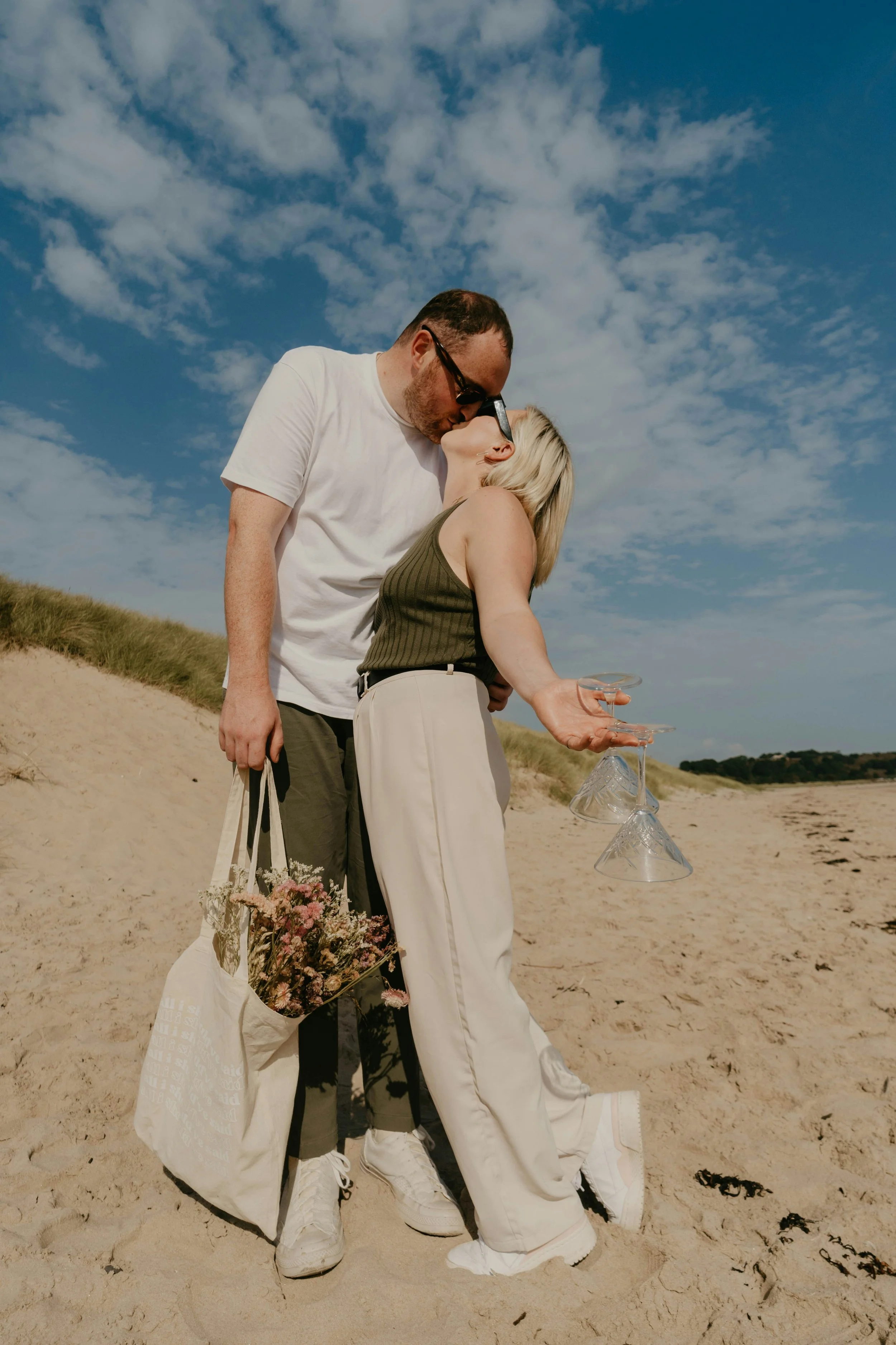 Couple sharing a kiss on the beach, holding dried flowers and empty martini glasses — representing celebration, affection, and partnership