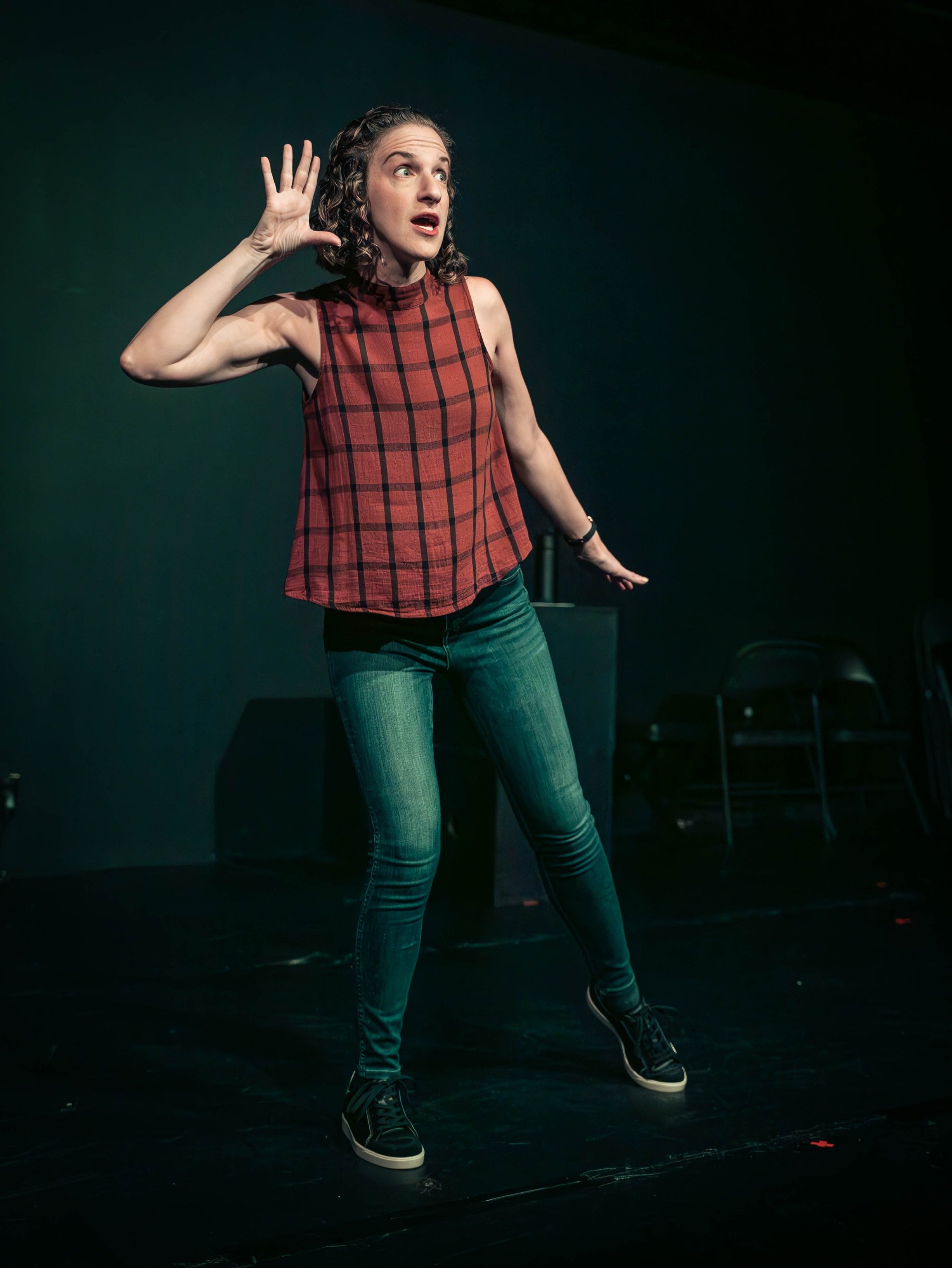 Woman with curly hair, wearing a red plaid sleeveless top, jeans, and sneakers, standing on a stage, holding her hand to her ear listening.