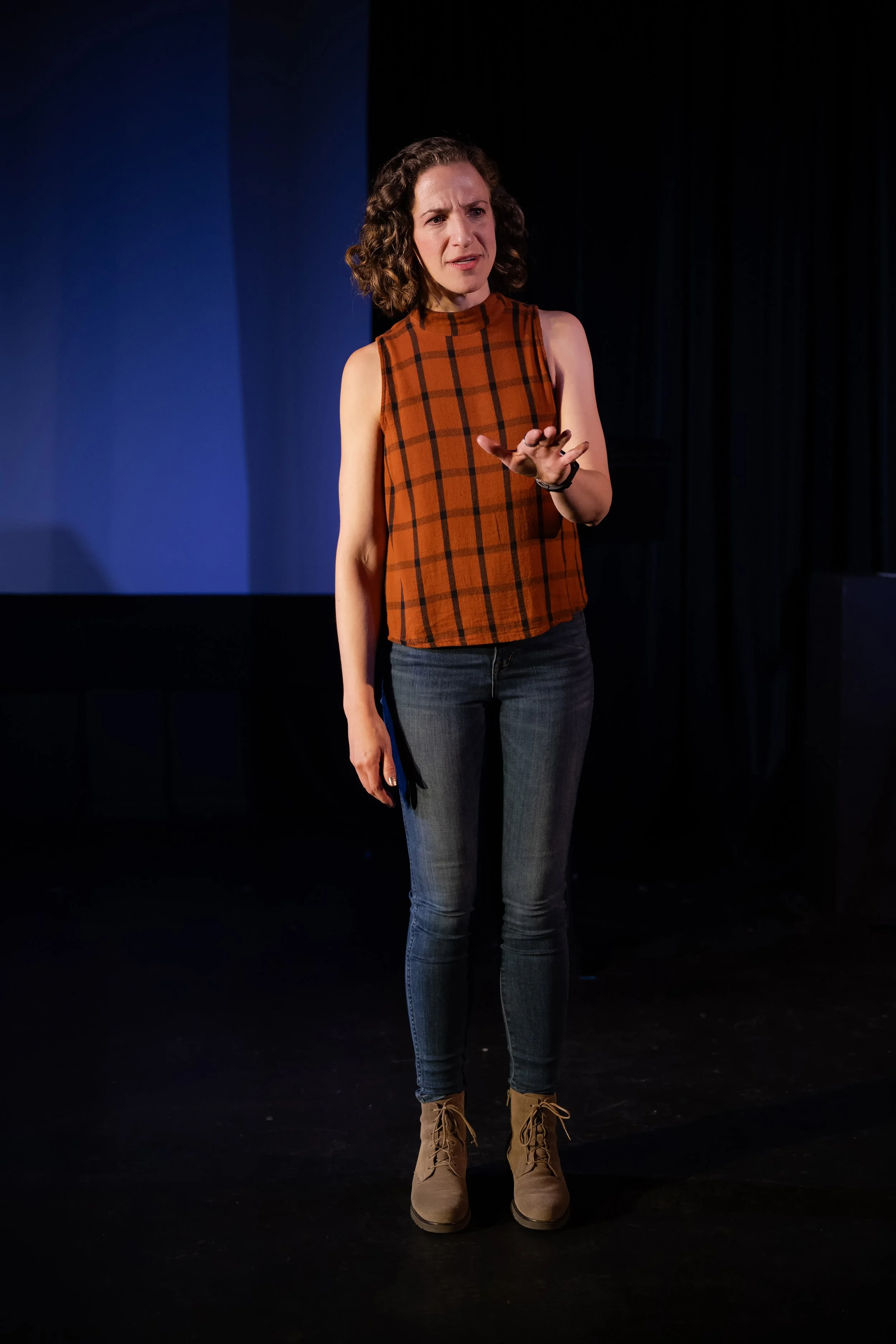 Woman with curly hair wearing a sleeveless orange and black plaid top, blue jeans, and tan boots, standing on stage against a dark background.
