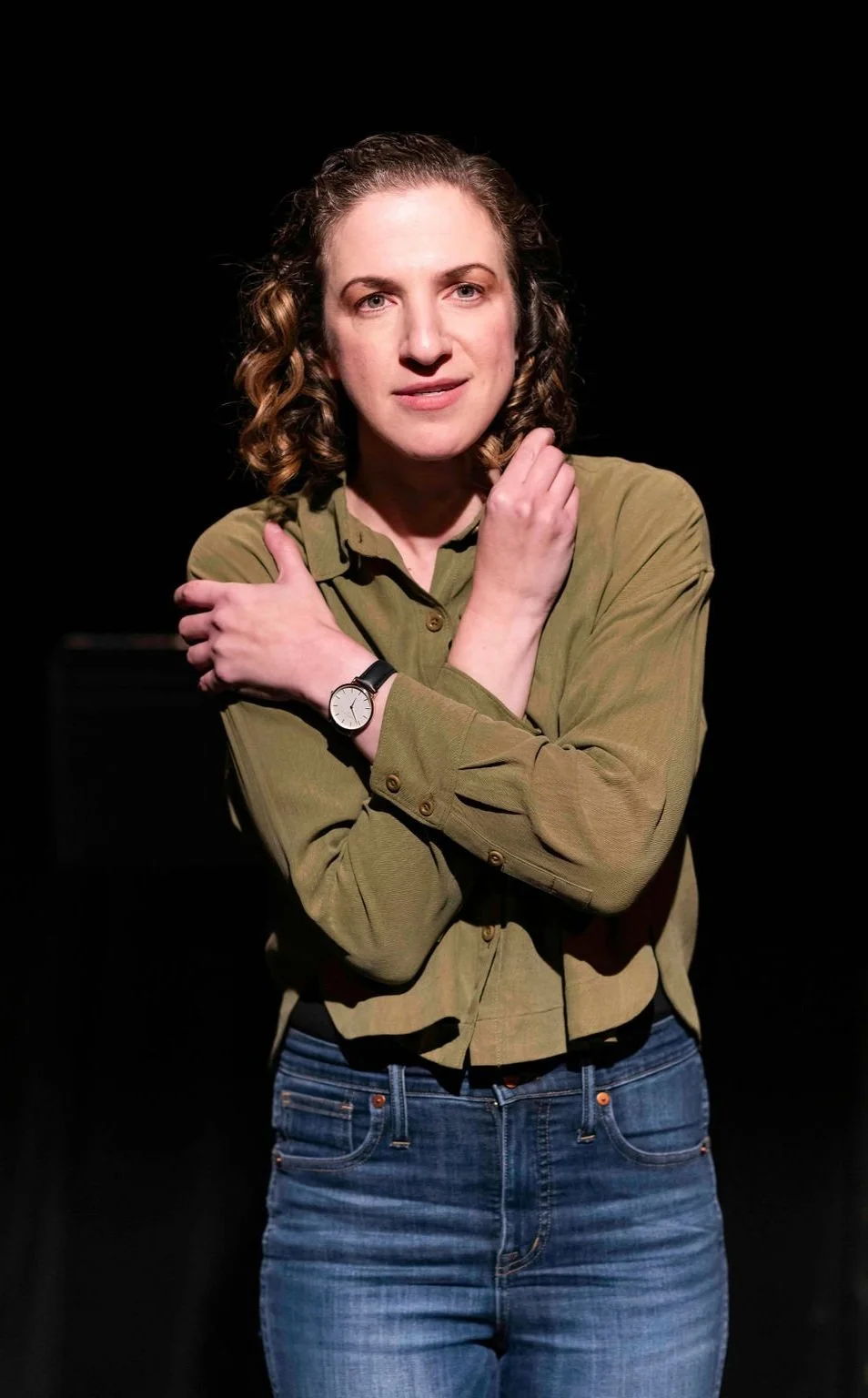 A woman with curly brown hair wearing an olive green shirt and jeans, standing on stage. She has her arms crossed, with her left hand touching her right shoulder and her right hand near her face.