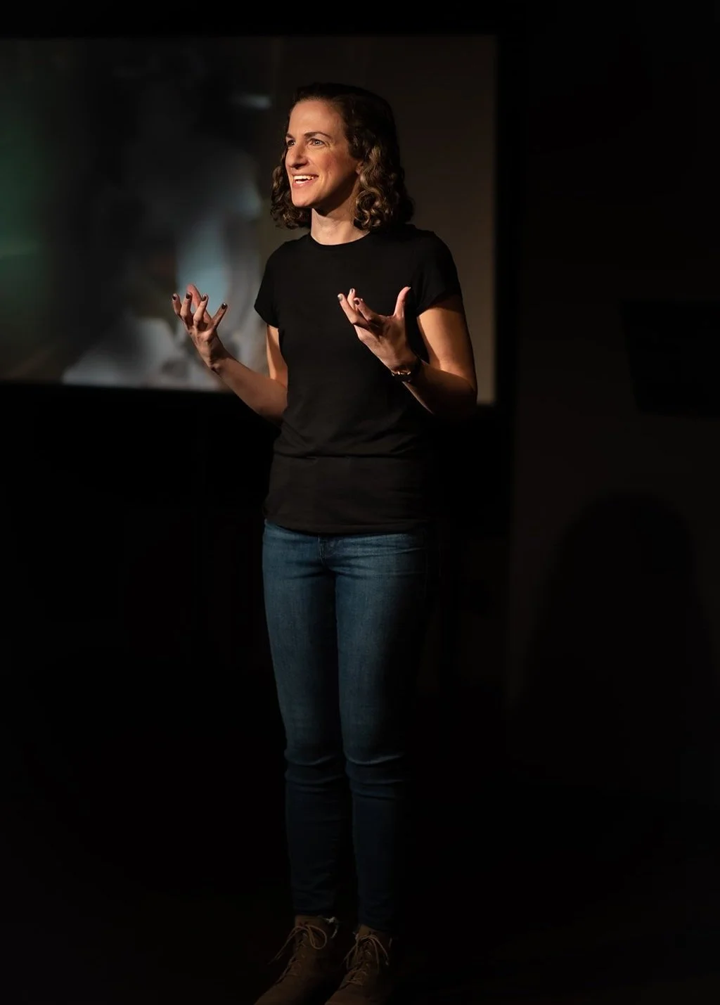 A woman with curly hair wearing a black T-shirt and jeans standing on stage speaking to the audience.