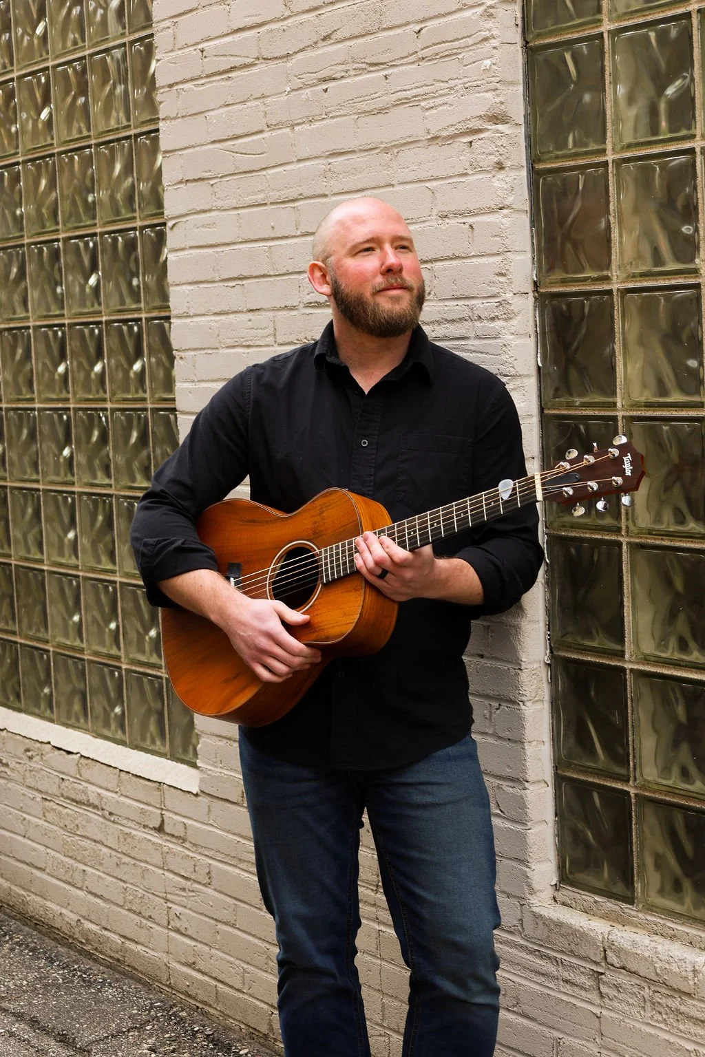 A man with a beard wearing a black shirt and jeans is holding an acoustic guitar and standing against a brick wall with glass blocks.