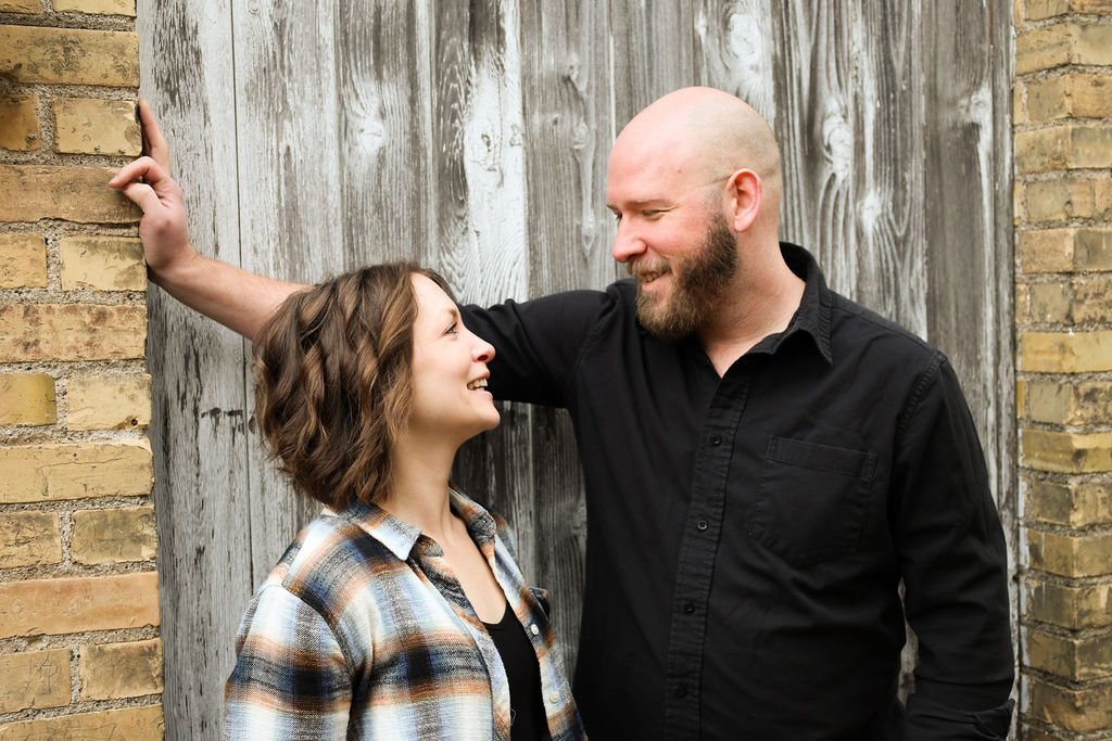 A man and woman smiling at each other, standing outdoors near a brick and wooden fence.