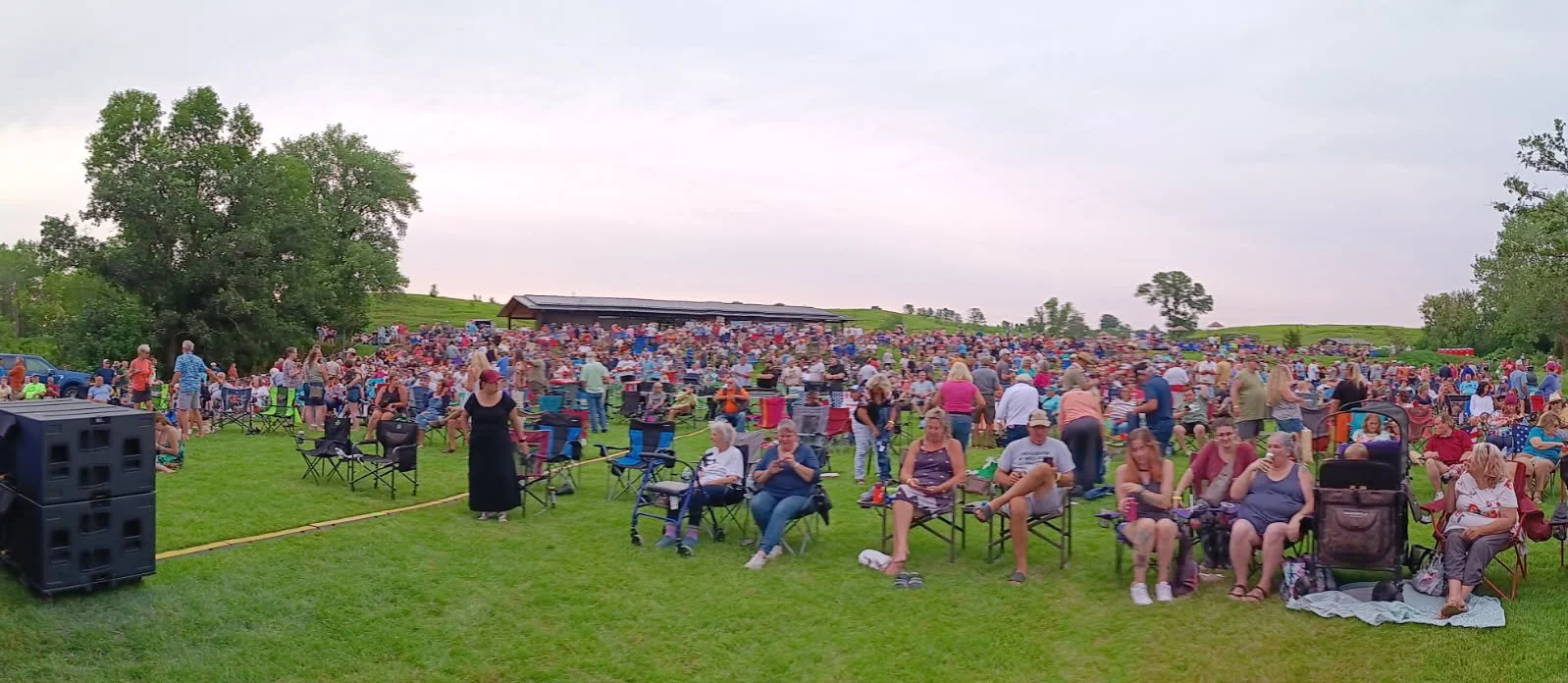 A large outdoor gathering where many people are sitting on lawn chairs and standing on a grassy field, with a structure in the background and trees on both sides, under a cloudy sky.