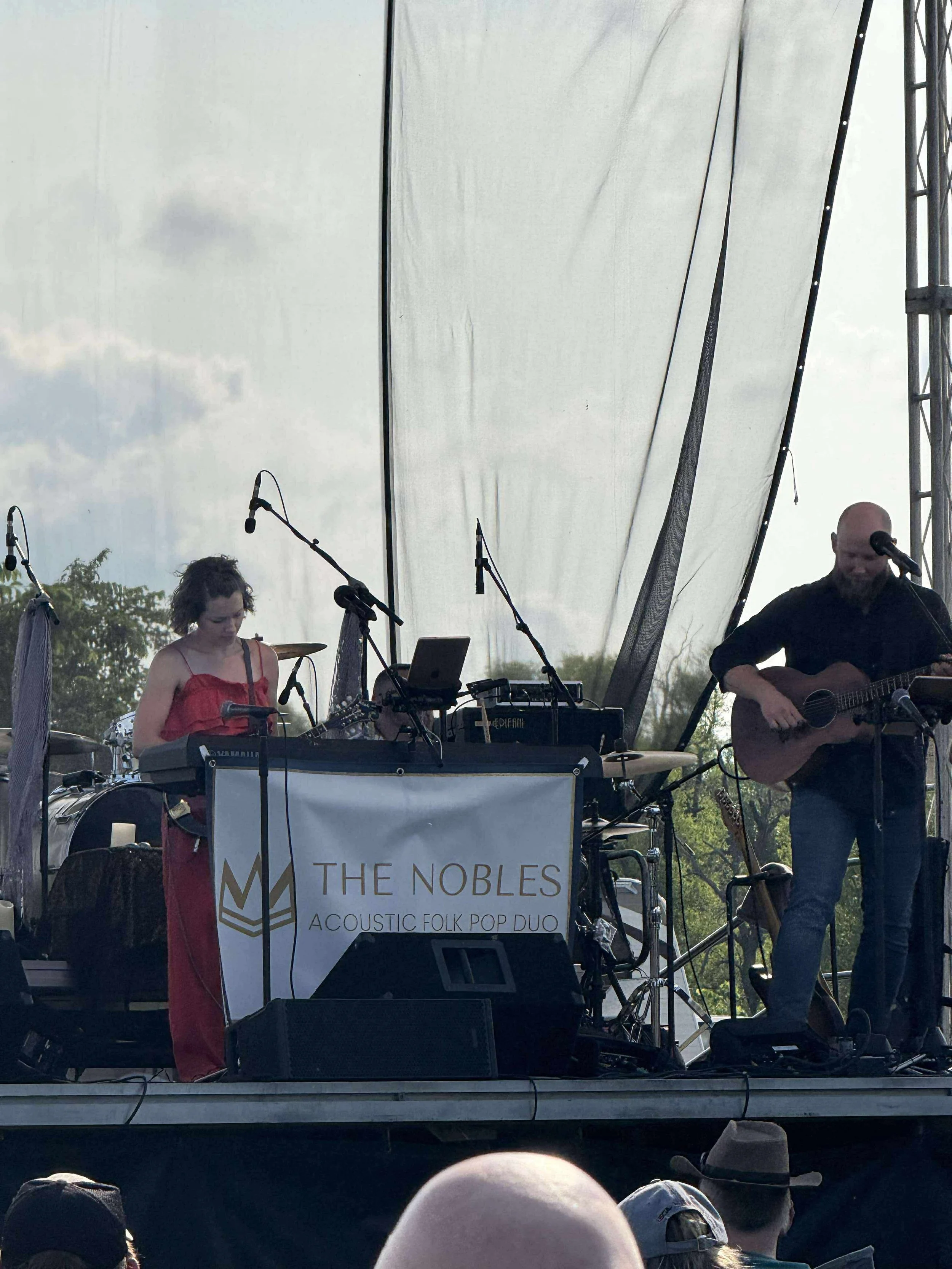 A live outdoor performance by a music duo called The Nobles, labeled as an acoustic folk pop duo, with two band members on stage. One woman playing keyboard and the other man playing guitar, with microphones and sound equipment in front of them. Audi