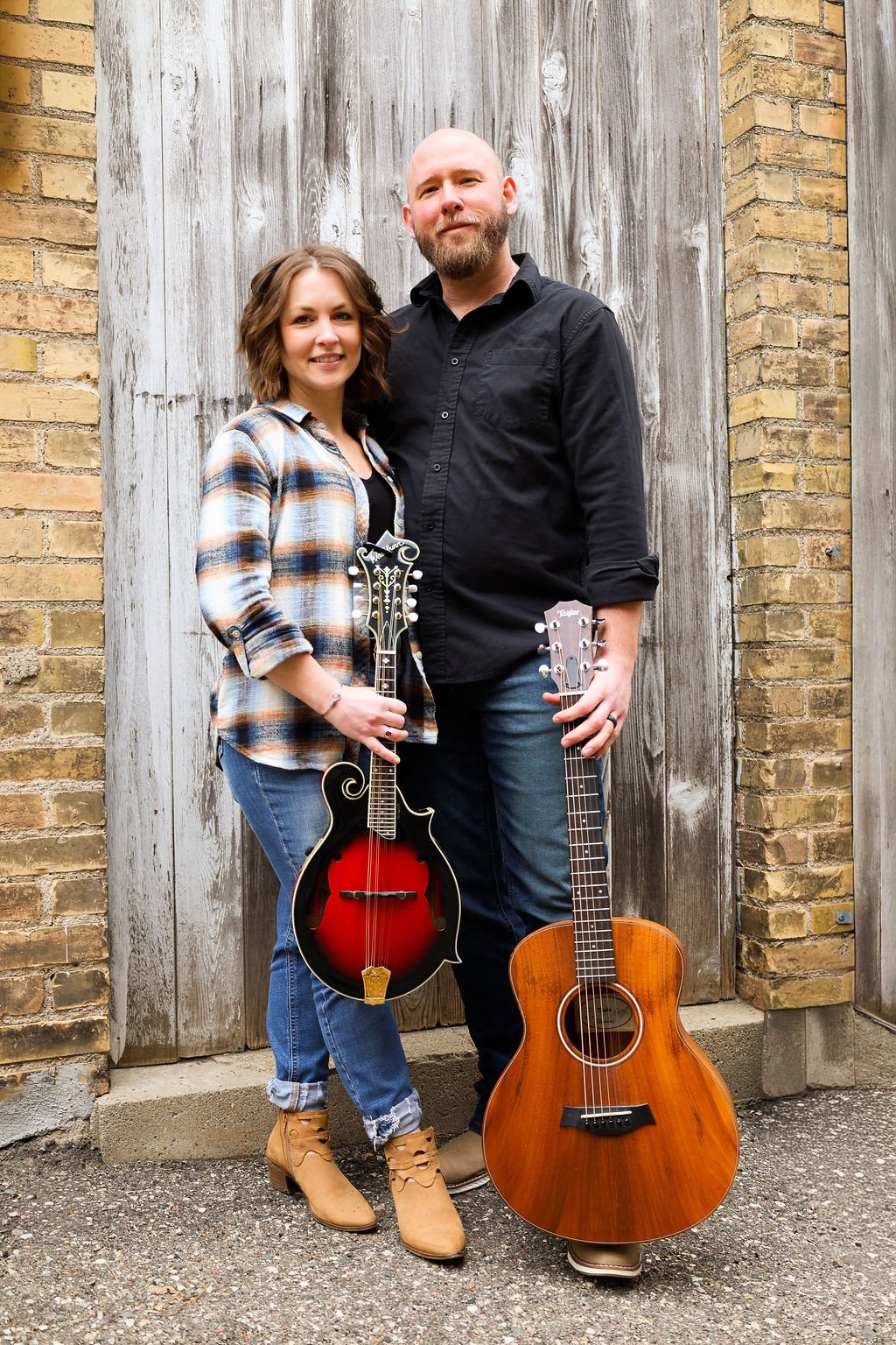 A man and woman standing outdoors against a wooden and brick wall, holding musical instruments: a mandolin and an acoustic guitar.