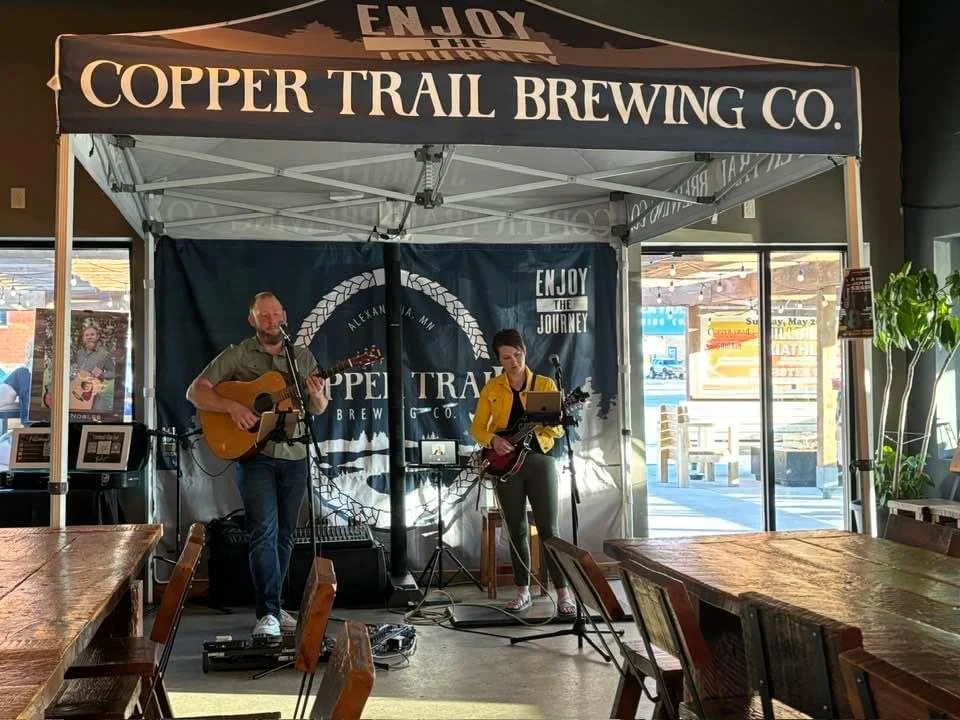 A live musical performance by a man and woman playing guitars on a small stage inside Copper Trail Brewing Co. The stage has a blue backdrop with the brewery's logo. The venue has wooden tables and chairs, with sunlight shining through large windows.