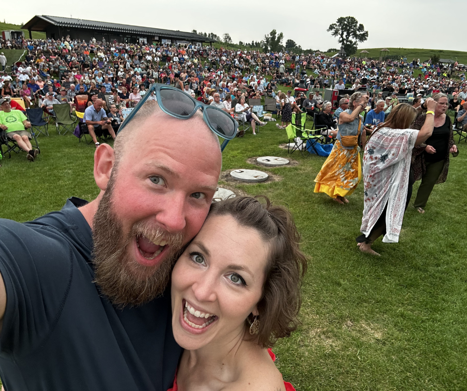 A man and woman taking a selfie at an outdoor concert with a large crowd in the background.