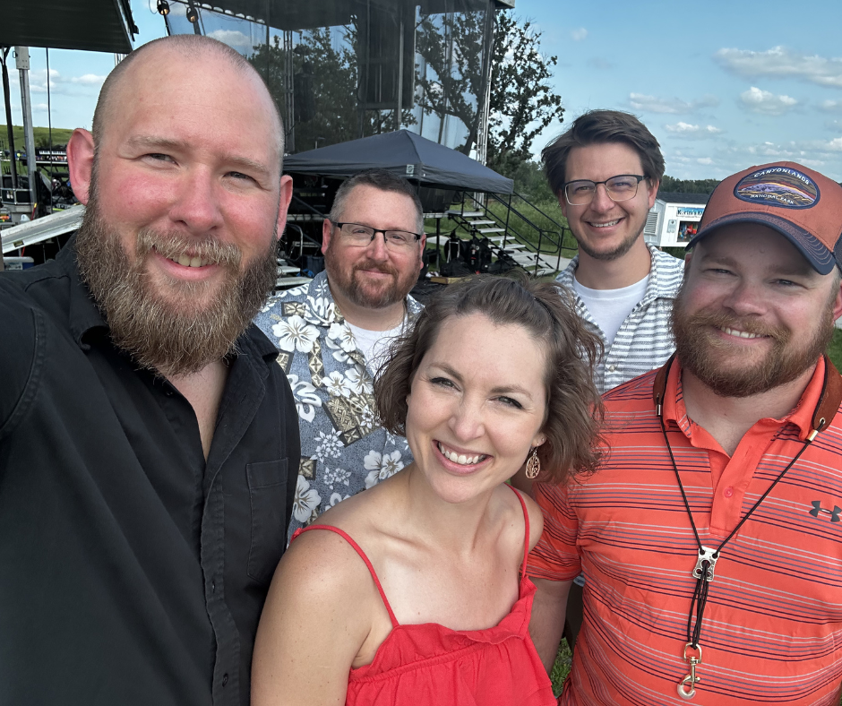 Group of five smiling people outdoors in front of a building with glass windows, on a grassy area with trees and a partly cloudy sky.