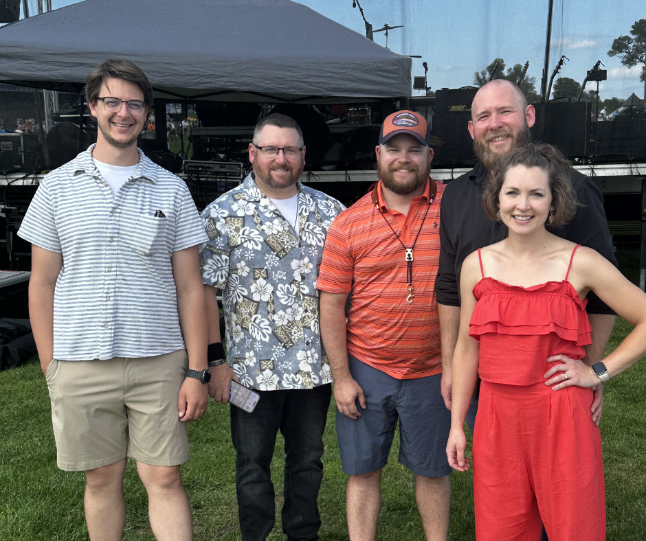 Group of five people enjoying outdoor event with stage and equipment in background
