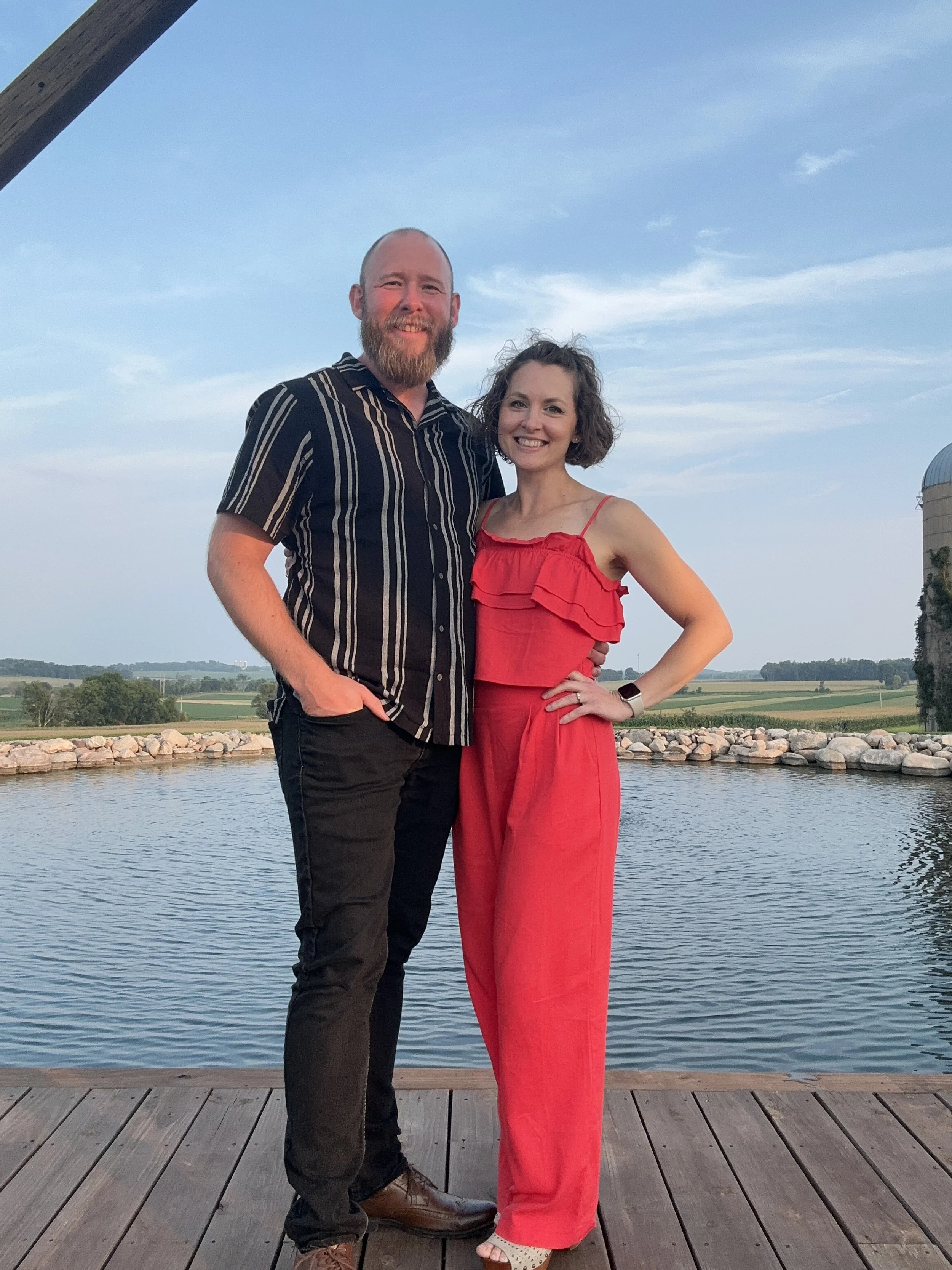 A smiling man and woman stand together on a wooden dock by a body of water, with a blue sky and distant landscape in the background. The man has a beard and is wearing a black striped shirt and black pants, while the woman has curly hair and is weari