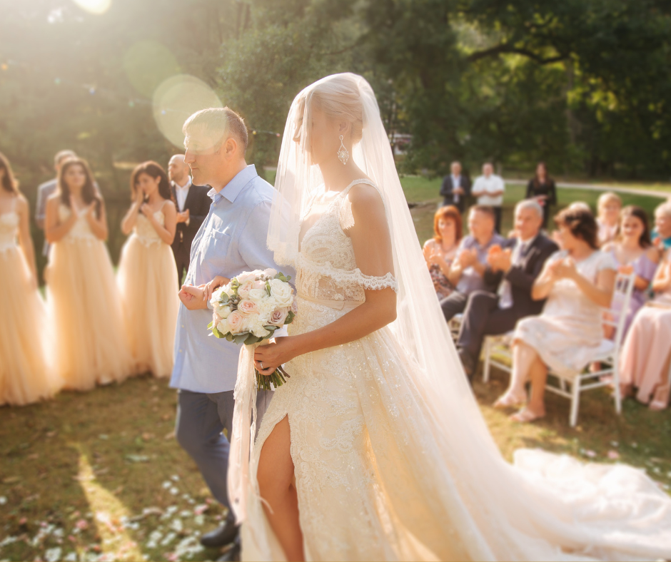 A bride in a lace wedding gown holding a bouquet of roses and a groom walking down the aisle outdoors with wedding guests seated and standing, sunlight shining through trees.