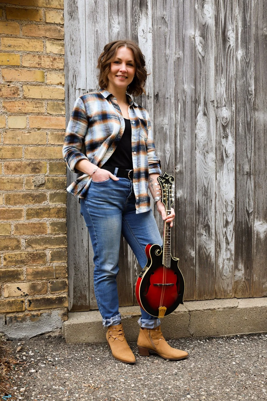A young woman with wavy brown hair, wearing a plaid shirt, black top, distressed blue jeans, and tan ankle boots, is standing outdoors against a background of brick and wooden panels. She is holding a vintage-style mandolin and smiling at the camera.