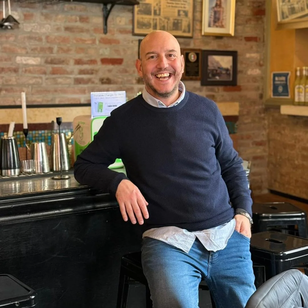 A smiling man with a beard and bald head, wearing a navy blue sweater over a white shirt and jeans, standing in a cozy restaurant or café with brick walls, framed pictures, and bottles on shelves in the background.