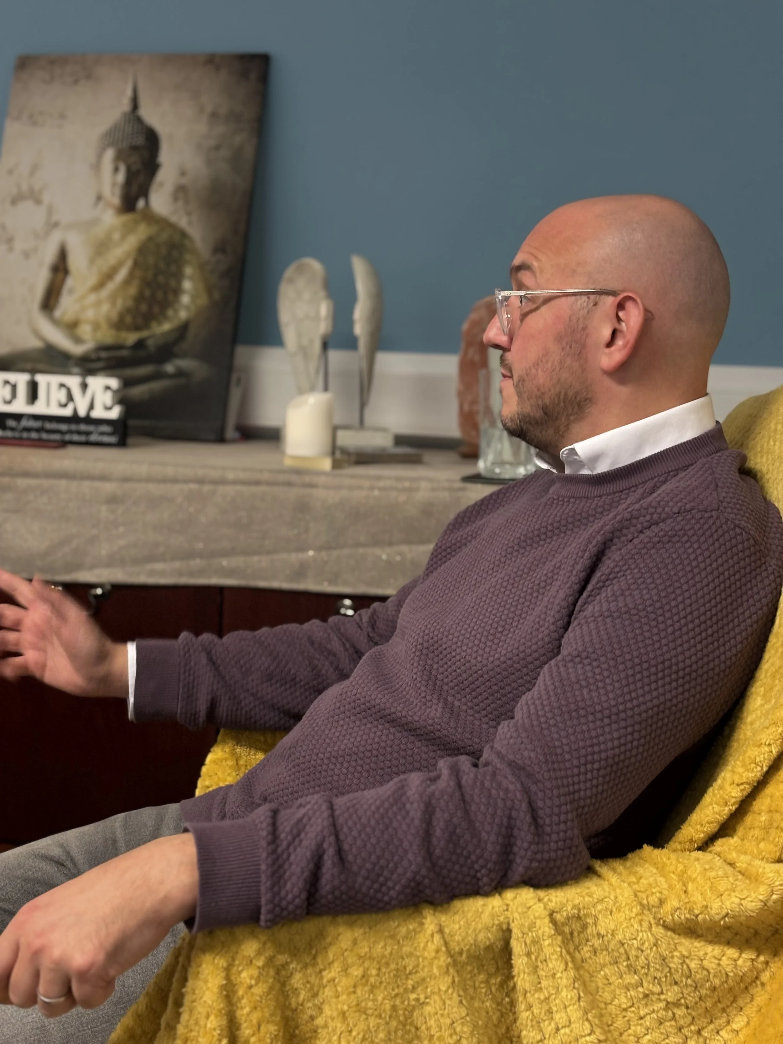 A man (Pietro Politano) with glasses and a bald head sitting in a yellow chair, in profile view. Behind him, a sideboard with a multi-panel Buddha painting, decorative angel wings, candles, and books on top. The wall behind is painted blue.