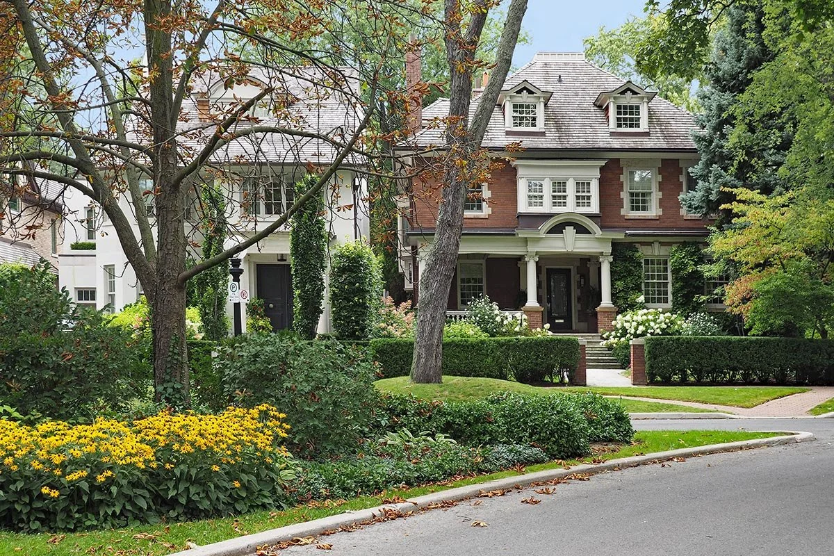A large, historic brick house with a covered porch, gabled roof, dormer windows, and a front yard with trees, bushes, and yellow flowers.