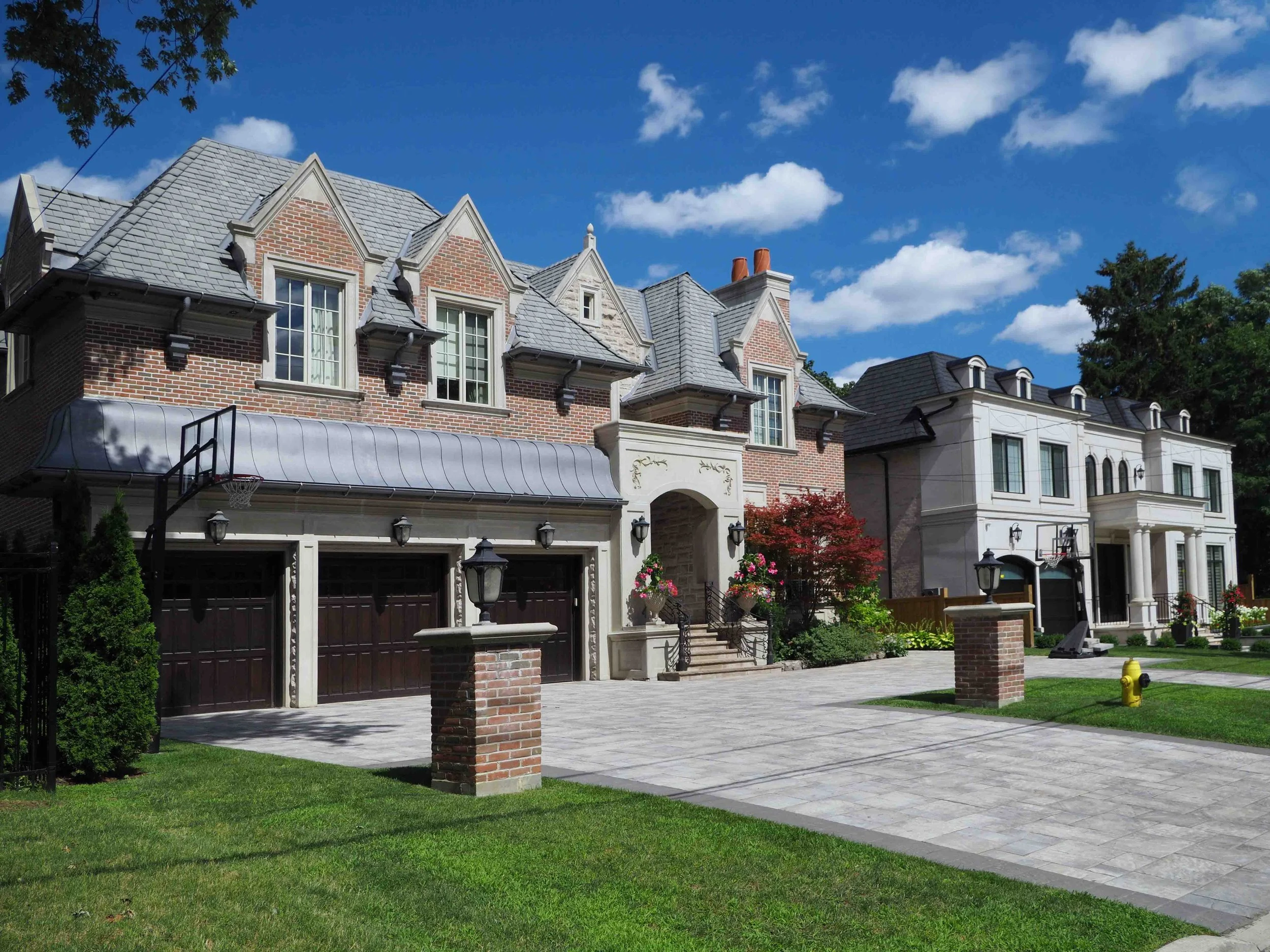 A large, elegant brick house in Forest Hill, Toronto, with a gray roof, white trims, and multiple dormer windows. The front yard features a paved driveway, green lawn, stone steps, decorative street lamps, and colourful plants and trees.