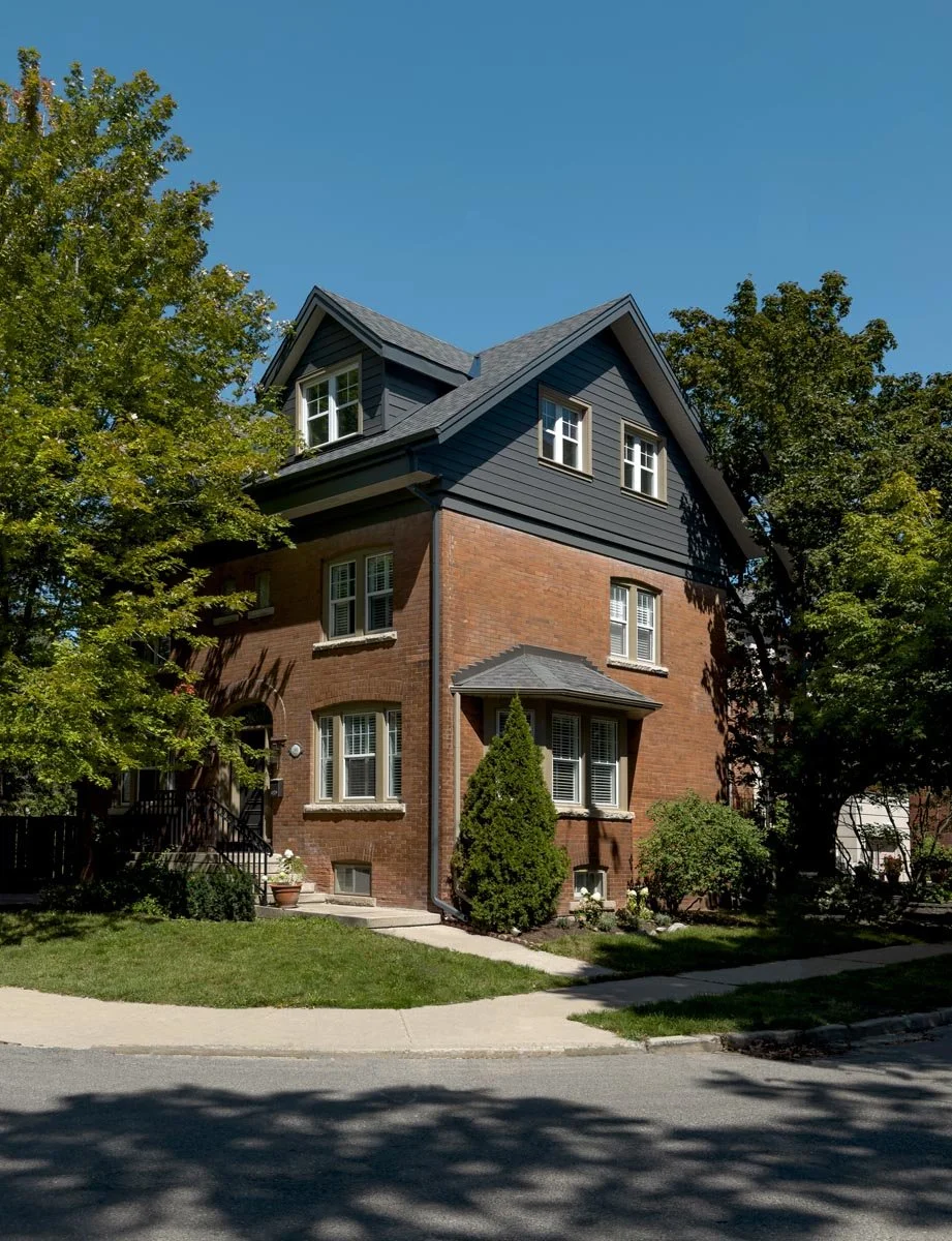 A three-story house with red brick on the lower levels and dark blue siding on the top floor. It has multiple white-framed windows, some with shutters, and is surrounded by green trees and a well-maintained lawn under a clear blue sky.