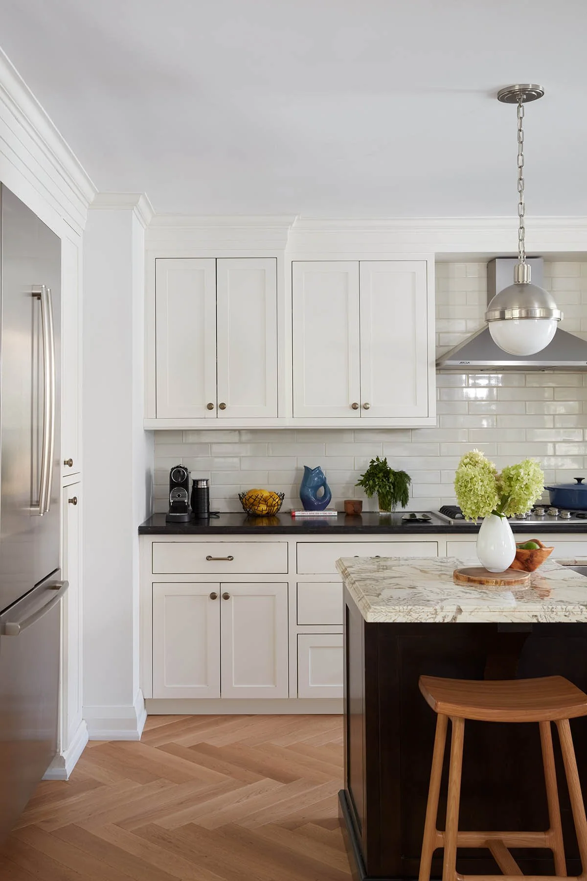 Home Renovation Services. Modern kitchen with white cabinets, black countertop, stainless steel appliances, and a marble-topped island with a wooden stool, decorated with a vase of green hydrangeas.
