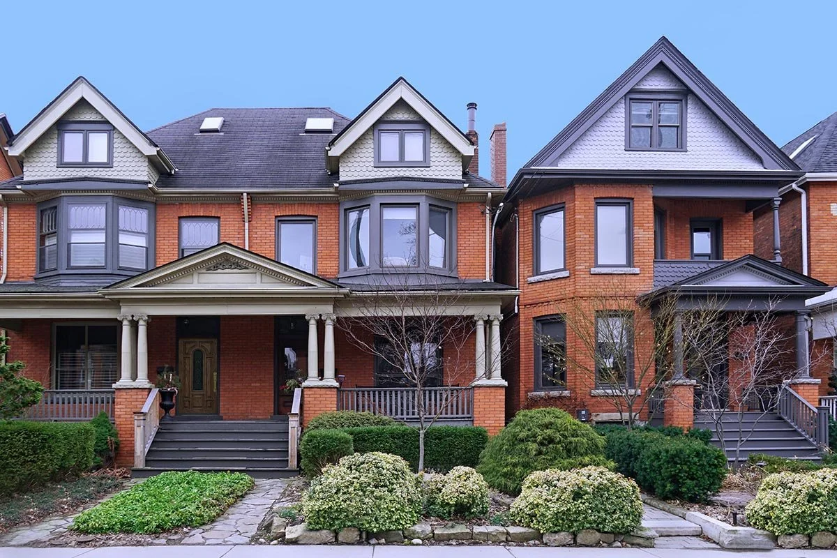A row of traditional Victorian-style houses with brick facades, gabled roofs, and front porches, surrounded by landscaped greenery and leafless trees.