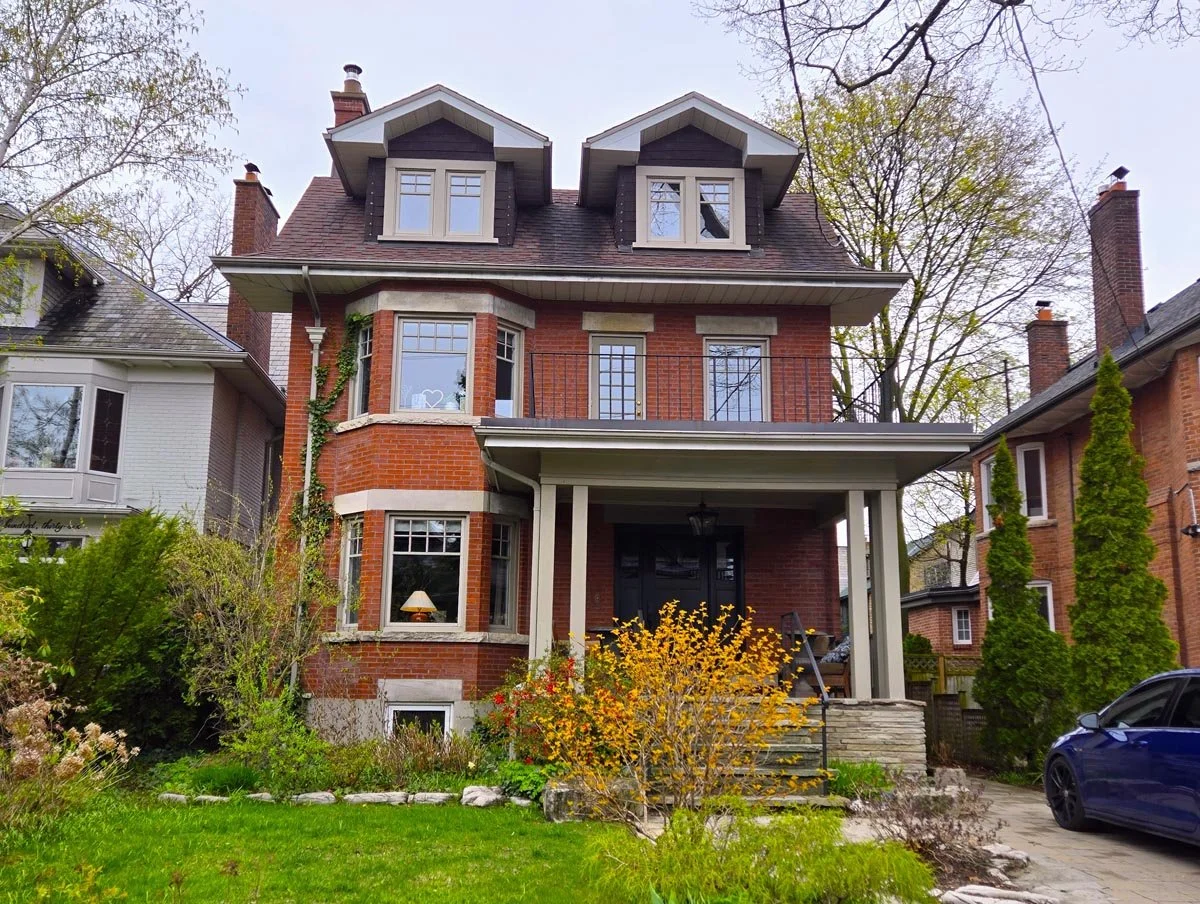 Front view of a three-story brick house with a covered porch, dormer windows, and a small front yard with plants and a yellow shrub.