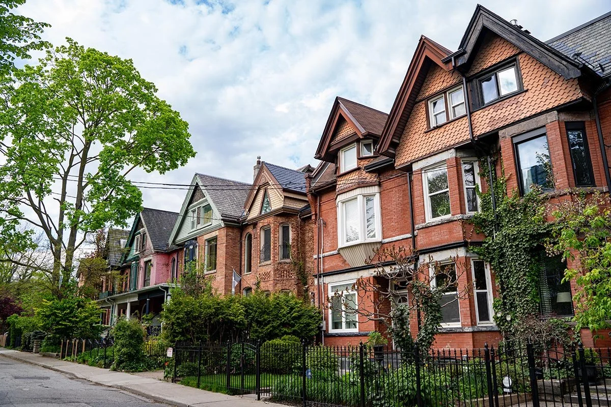 A row of historic brick houses with decorative gables, front porches, and trees lining the sidewalk.