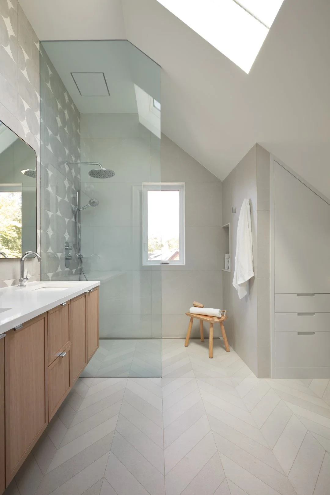 Bathroom renovation with light grey tile, natural woods and glass. Standing shower open to bathroom vanity and storage area.
