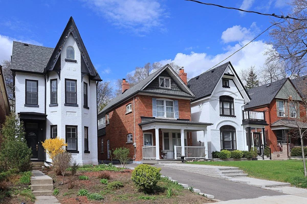 A row of three houses on a clear day with blue sky and white clouds, with a sidewalk and small garden in front.