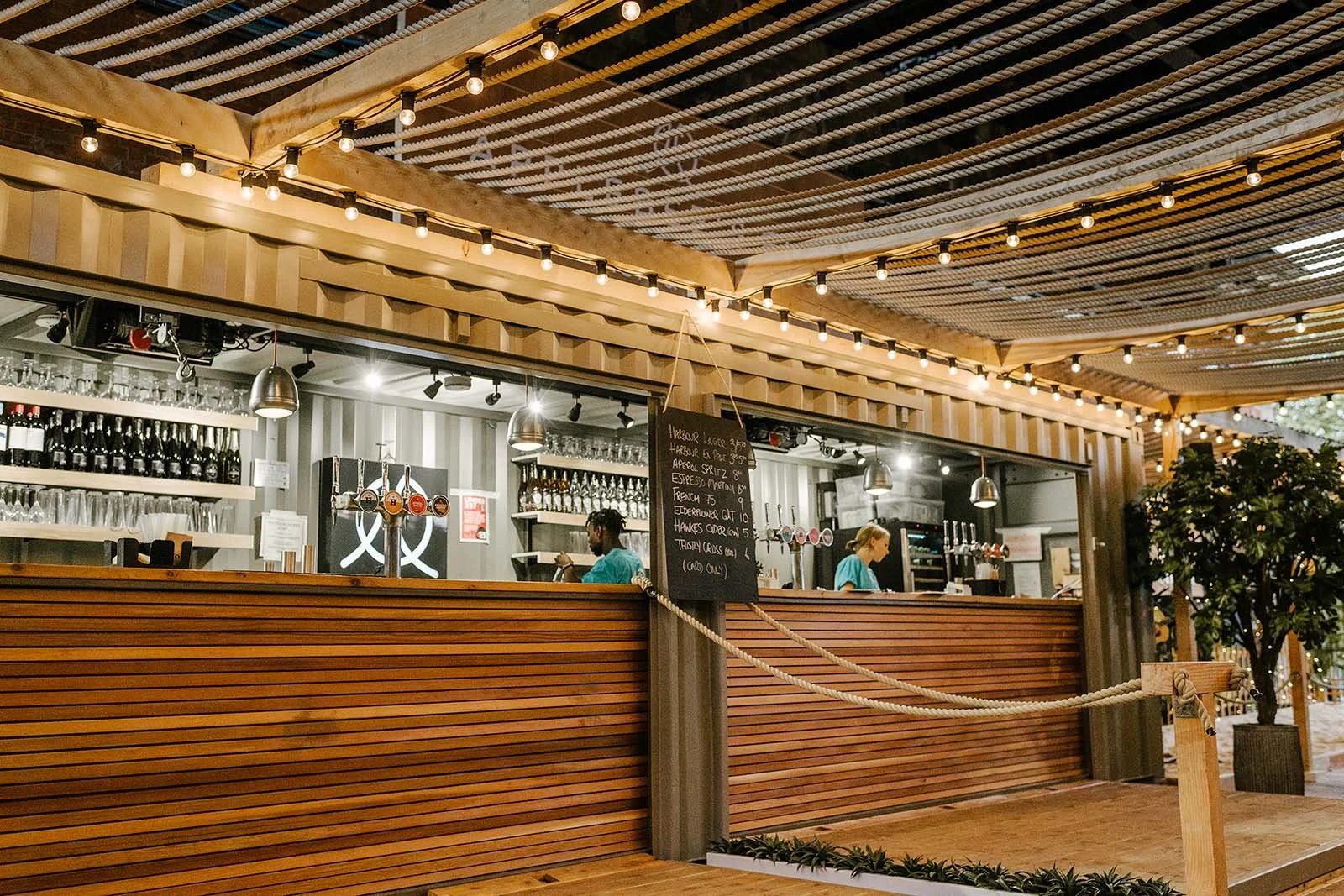 Inside a modern cafe with large glass windows showing outdoor water views, a woman on the phone, and a barista behind the counter with green decorative items on the wall.