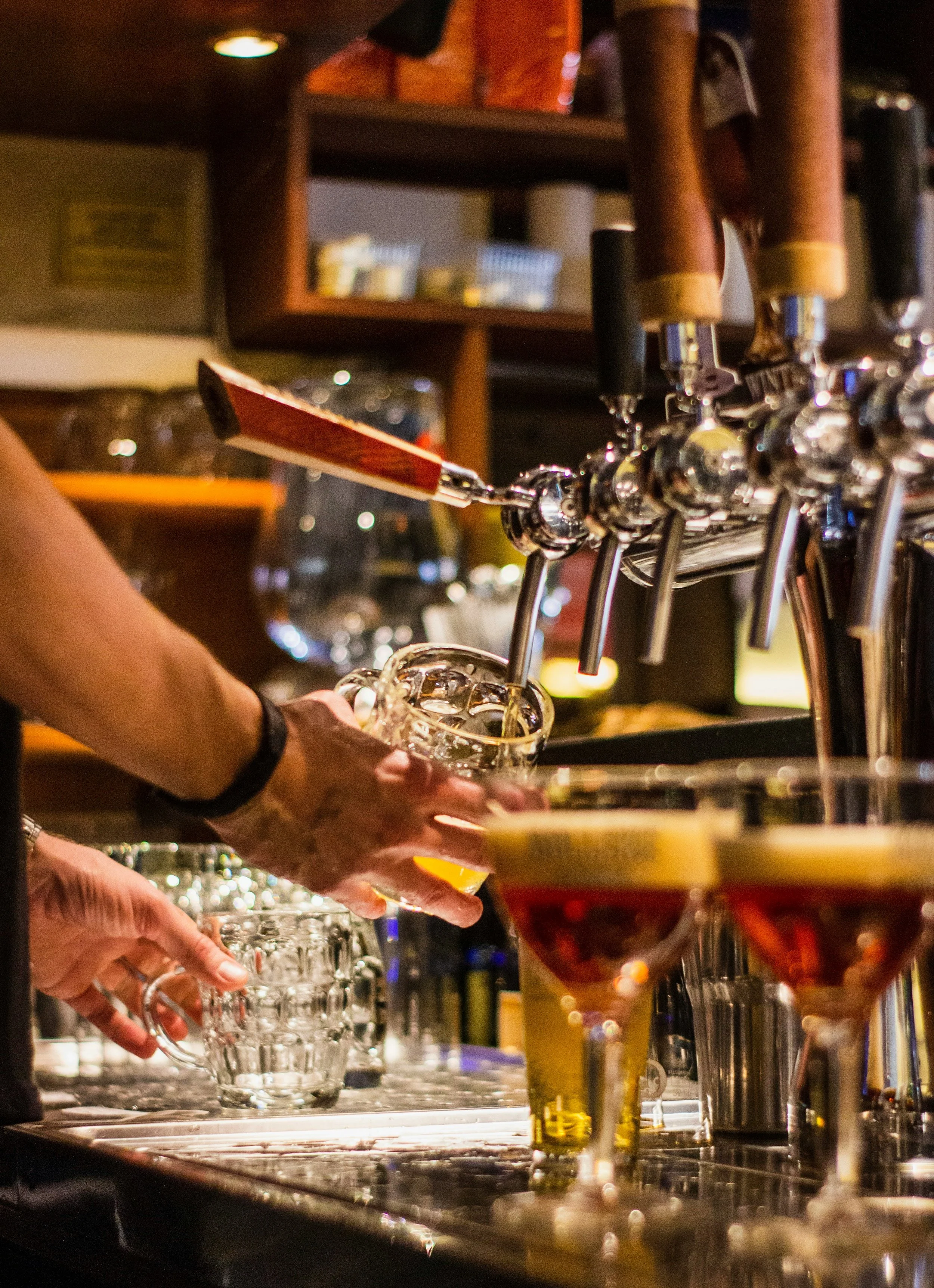 A bartender is pouring draft beer from tap handles into a glass, with two cocktails with garnishes in the foreground.