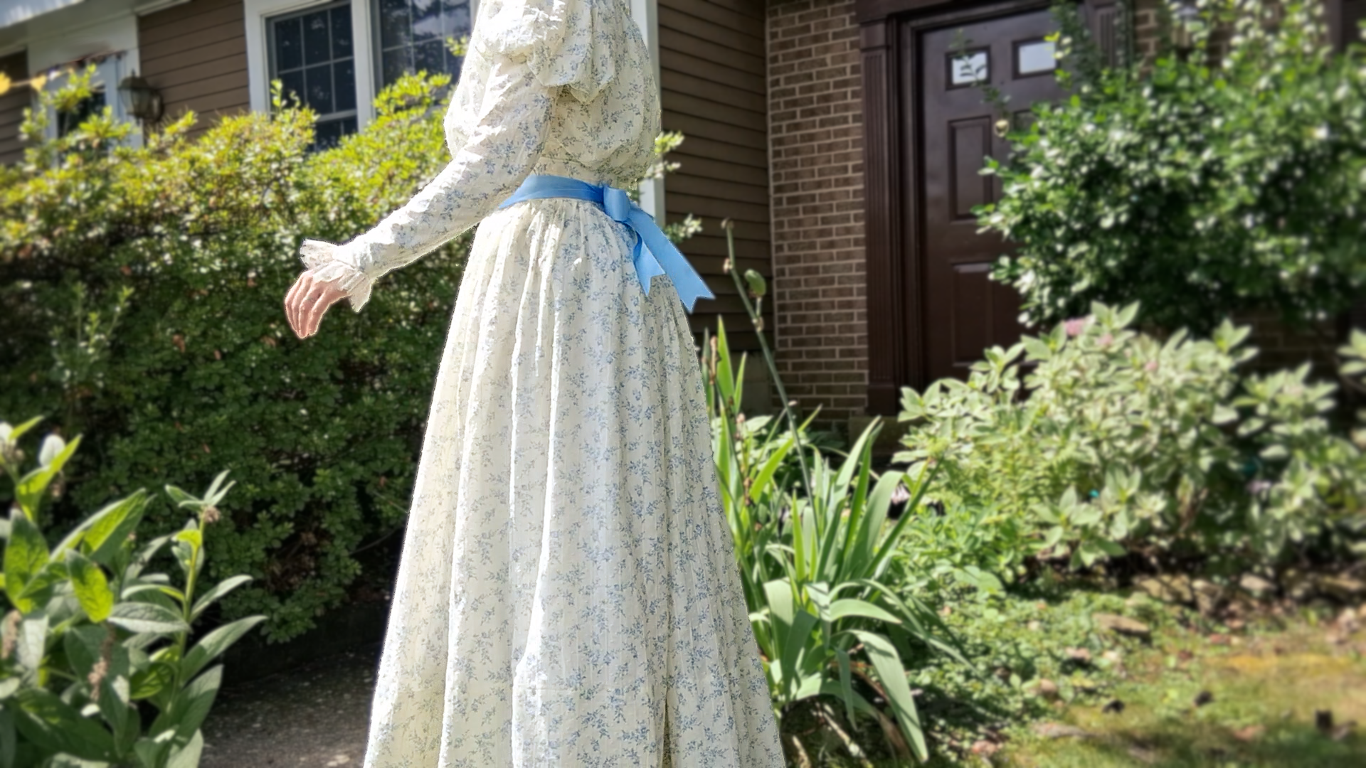 Side profile of Kyla wearing the completed 1890s Alice Dress, a cream historical gown with a delicate floral print and a bright blue waist ribbon tied in a bow at the back.