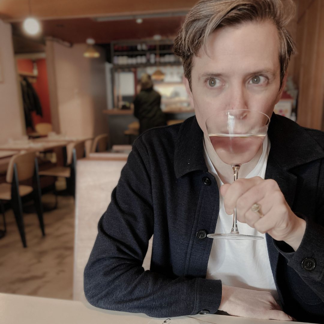 Anj Gierak, co-director of Bold Green Strategies. A man with short light brown hair, sitting at a table in a restaurant, holding a martini, bringing it up to his mouth. He wears a white t-shirt and a dark jacket, with a gold ring on his ring finger.
