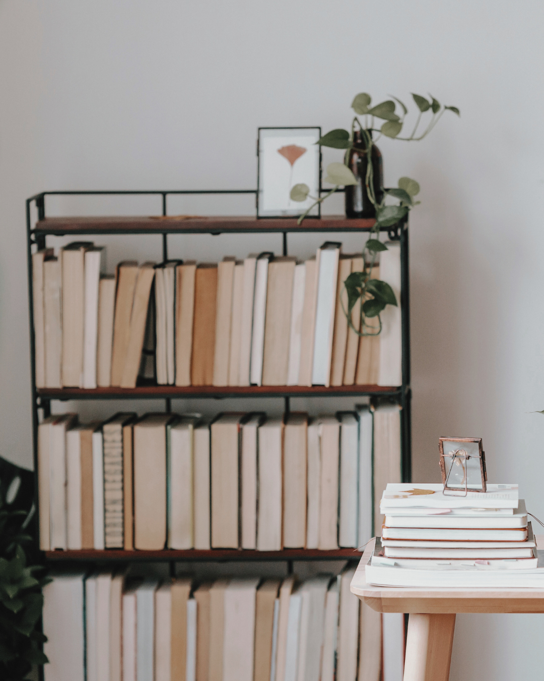 A black metal and wooden bookshelf filled with cream-colored books, with a small table holding a stack of books and framed photos, along with a plant on top of the bookshelf.