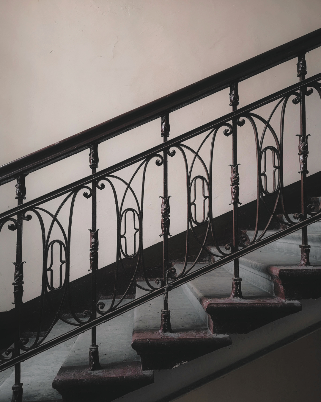 An ornate black iron railing on a narrow staircase with dark red steps, against a plain beige wall.