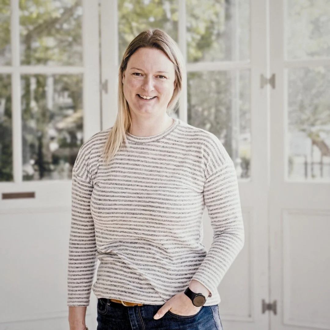 Cath Harris, marketing consultant. Woman smiling, wearing a striped shirt and jeans, standing indoors with large windows showing trees outside.