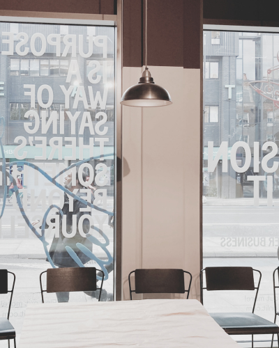 Interior of a modern cafe or coffee shop with large windows displaying text, a hanging pendant light, a white table, and black chairs.