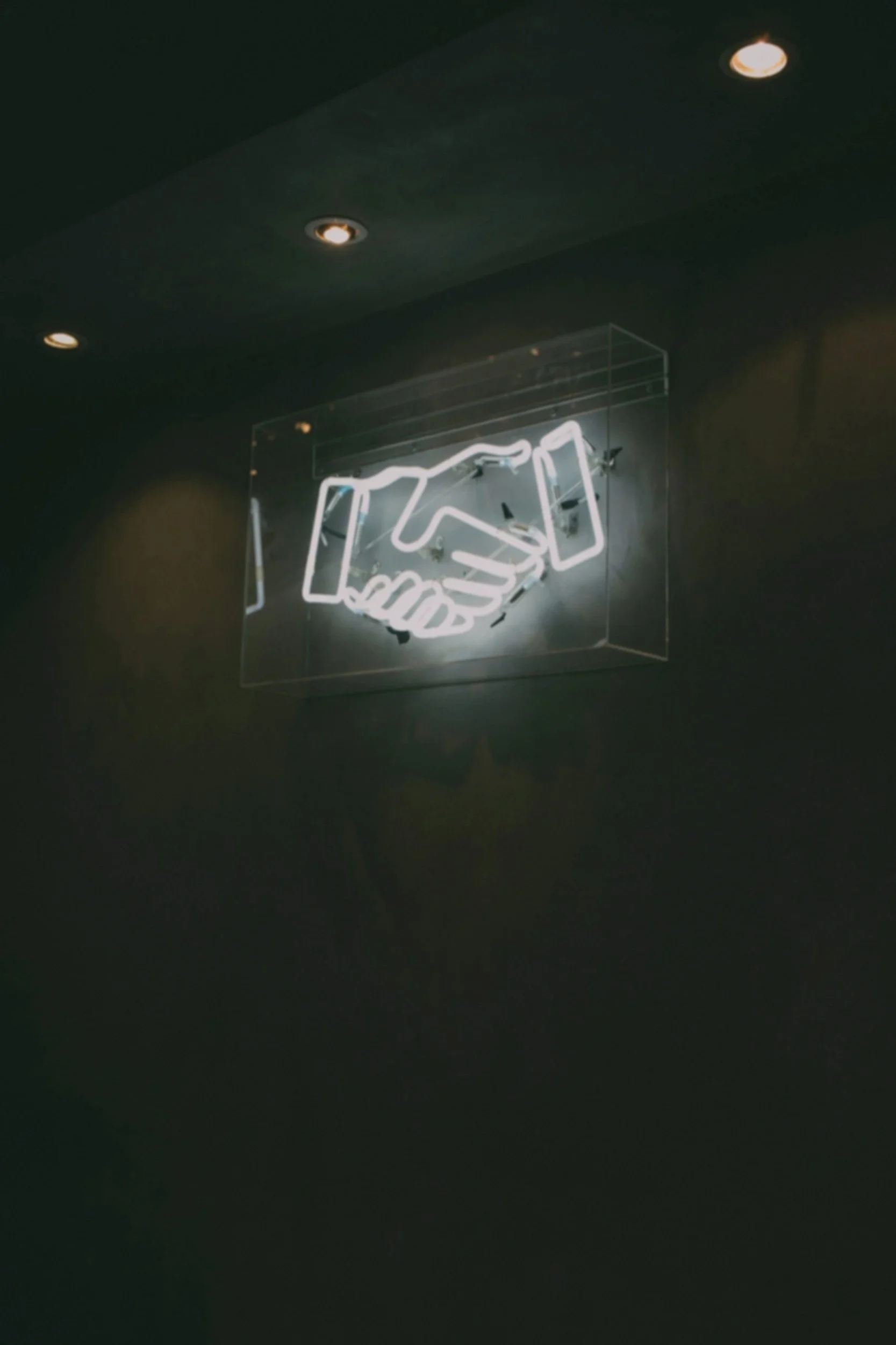A sign with a white neon handshake on a dark wall representing the opportunities sustainability and impact reporting provide in tenders, procurement, compliance, and maintaining partnerships.