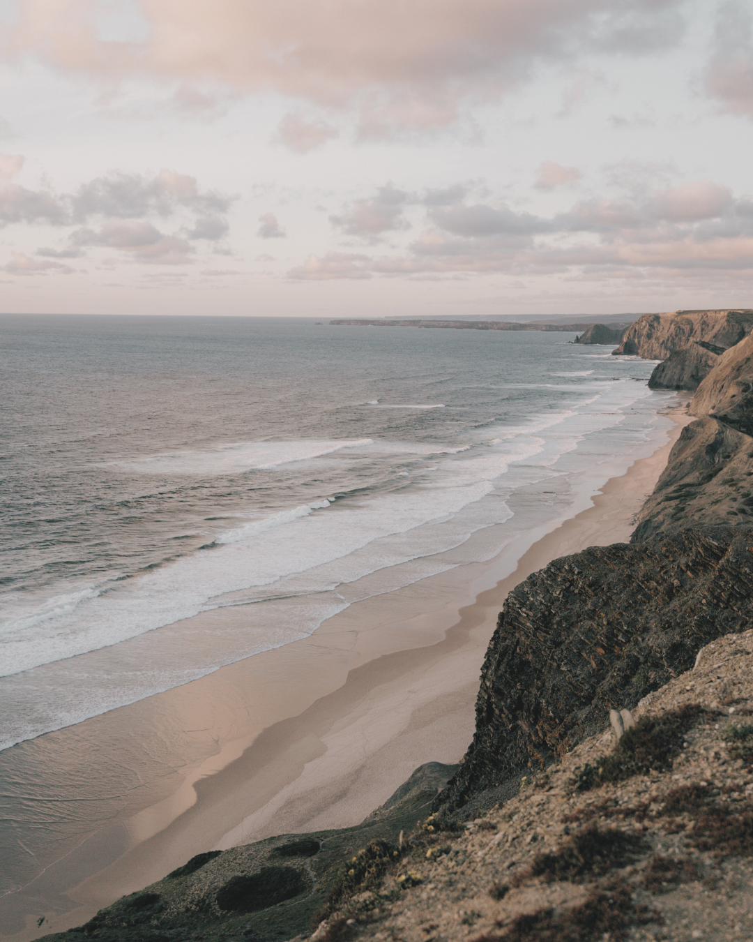 Cloudy sky over a coastal beach with waves crashing on the sandy shore and tall cliffs along the coast.
