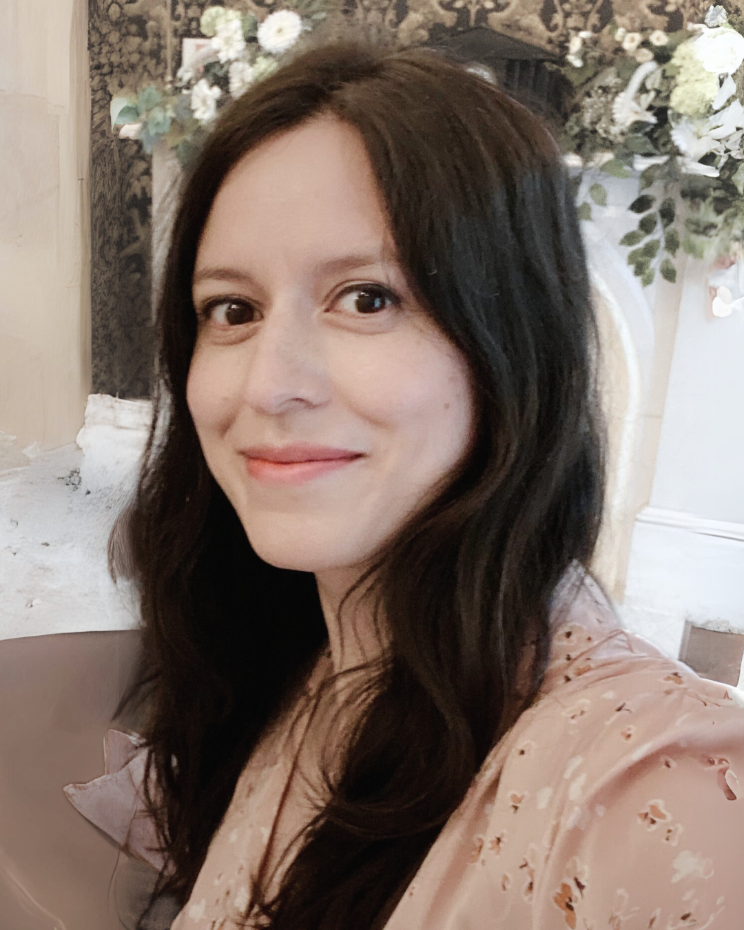 Lina Ahmed. A woman with long wavy dark hair smiling in front of floral arrangement.