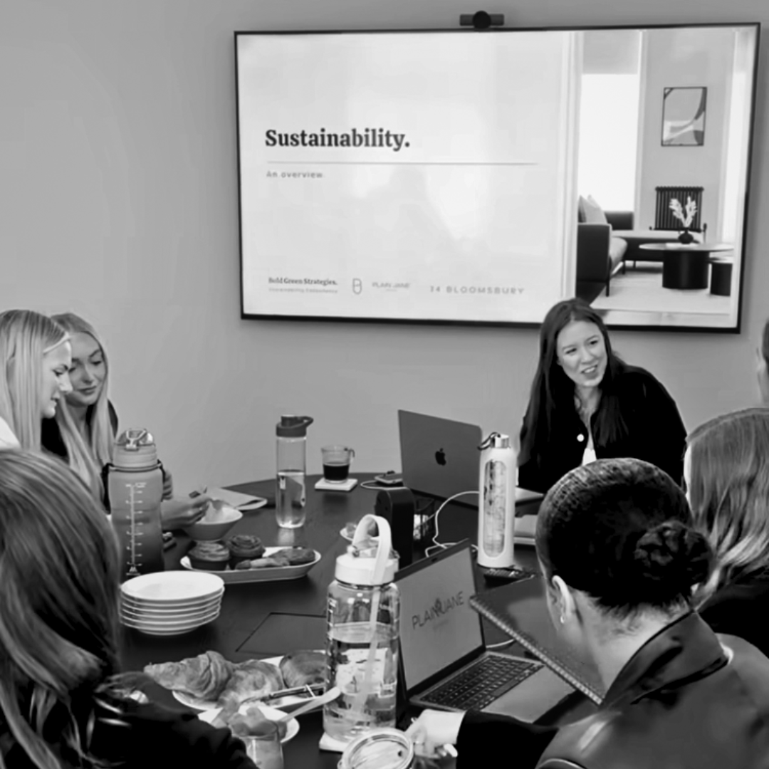 A group of women attending a presentation or meeting on sustainability in a conference room. There are laptops, water bottles, snacks, and a large screen displaying the presentation.