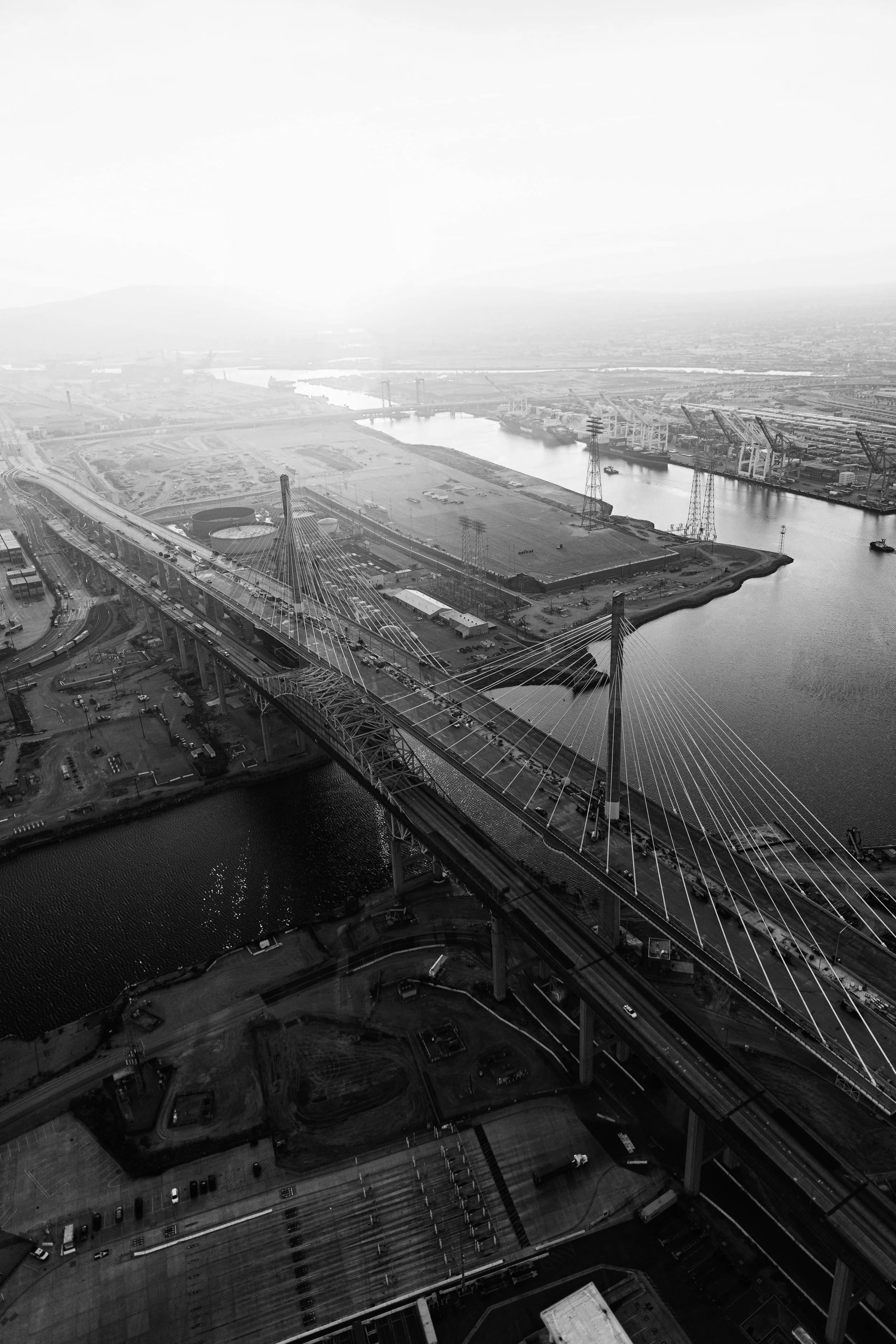 Aerial view of a cityscape featuring a large suspension bridge, industrial port with cranes along the water, and roads with vehicles. The landscape is flat with some distant mountains or hills.