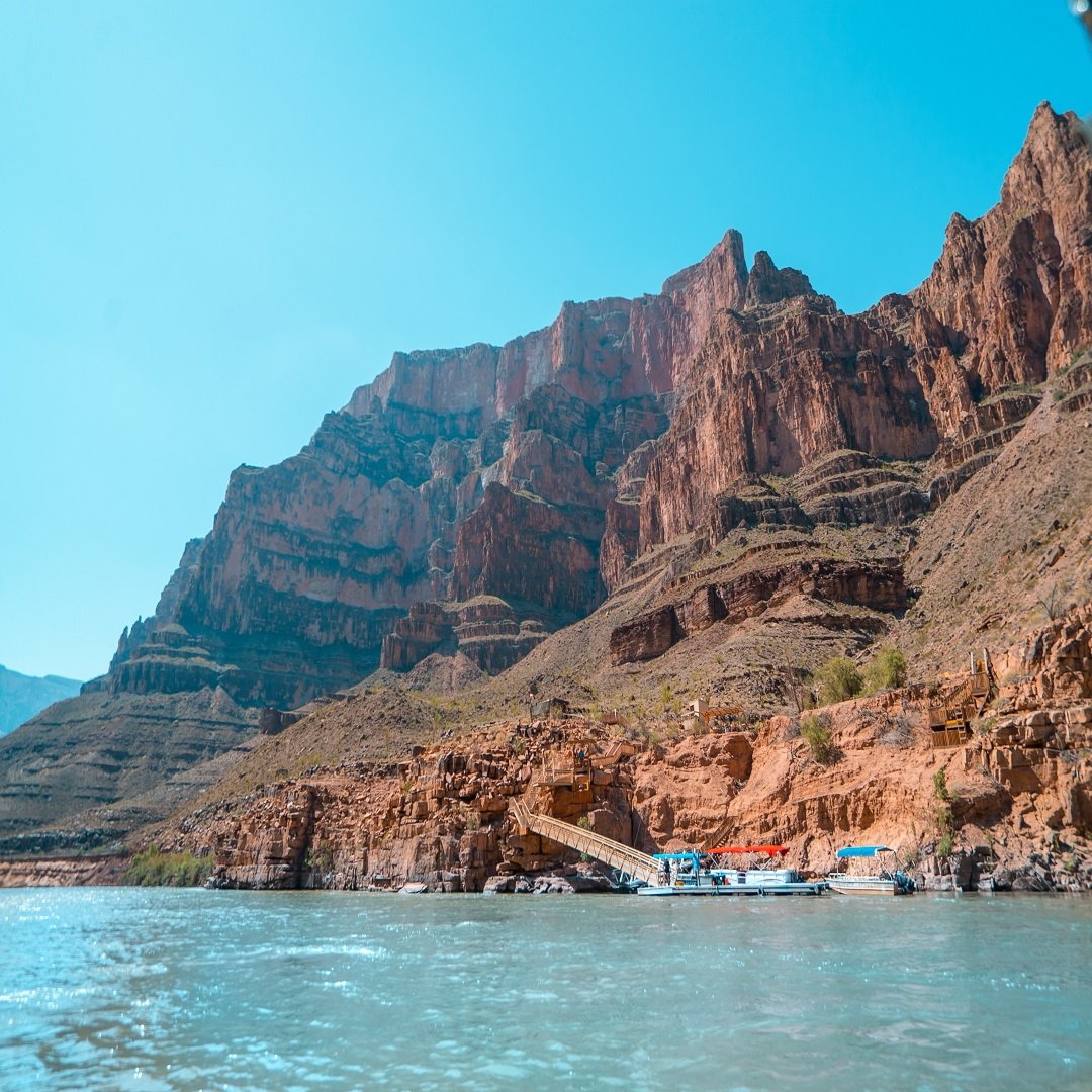 View of the Grand Canyon with steep rocky cliffs and layers of reddish-brown rock formations towering over the Colorado River at the bottom, under a clear blue sky.