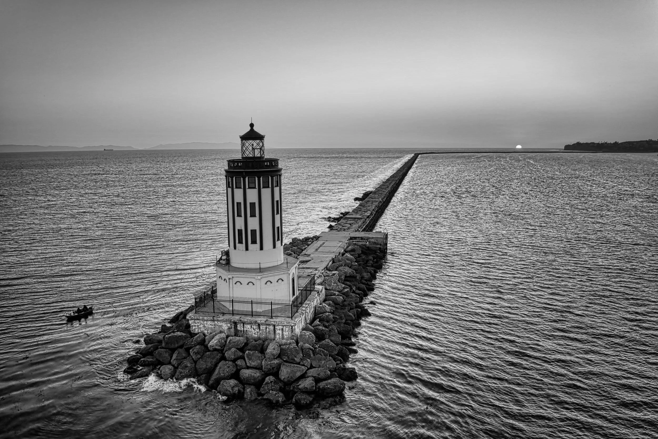 A black and white photo of a lighthouse on a rocky pier extending into the water, with the sun setting on the horizon.