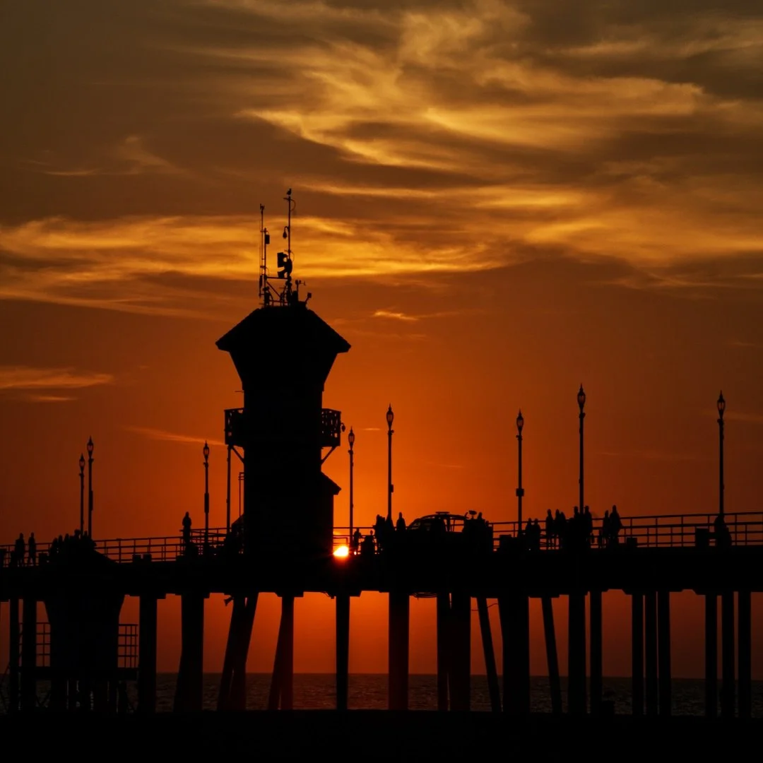 Silhouette of a pier with a lighthouse and people during a vibrant sunset with clouds in the sky.