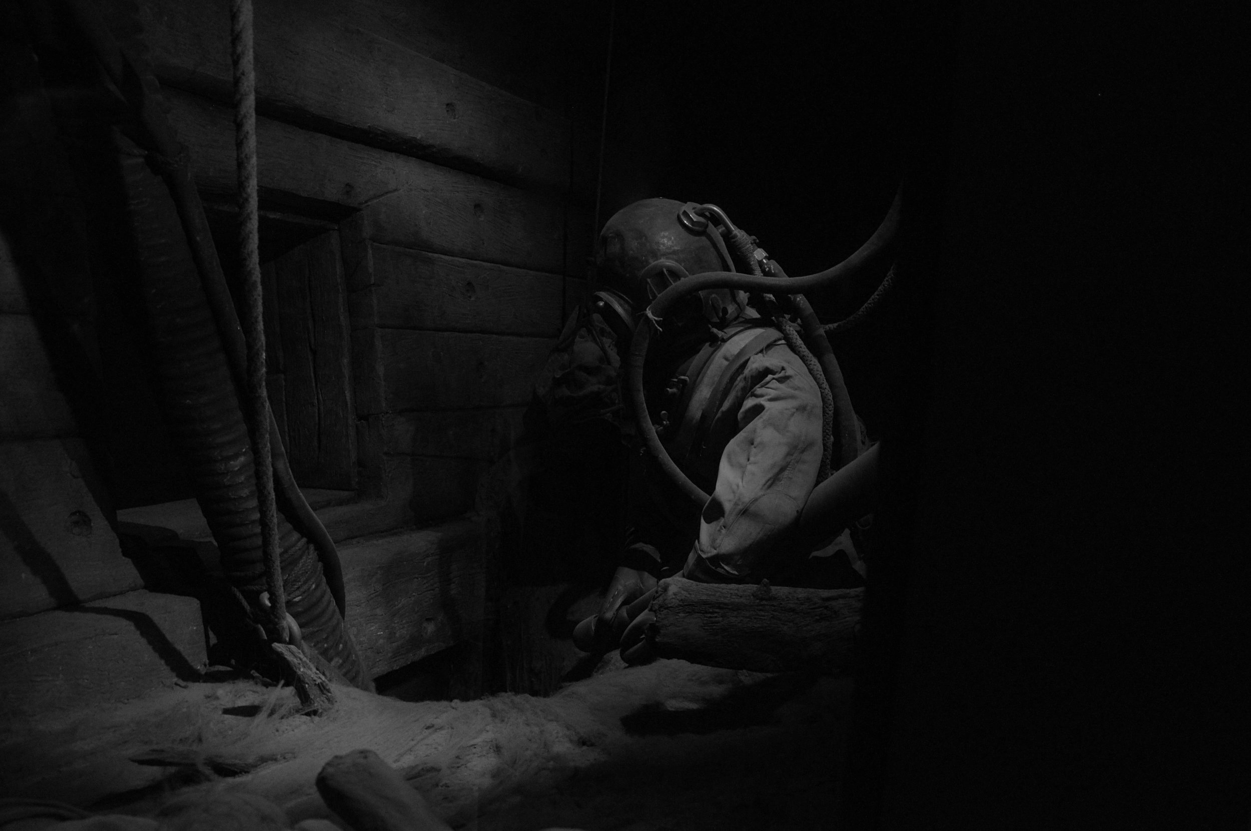 A miner in a dark, enclosed space wearing a helmet and a protective suit, surrounded by pipes and wooden beams.