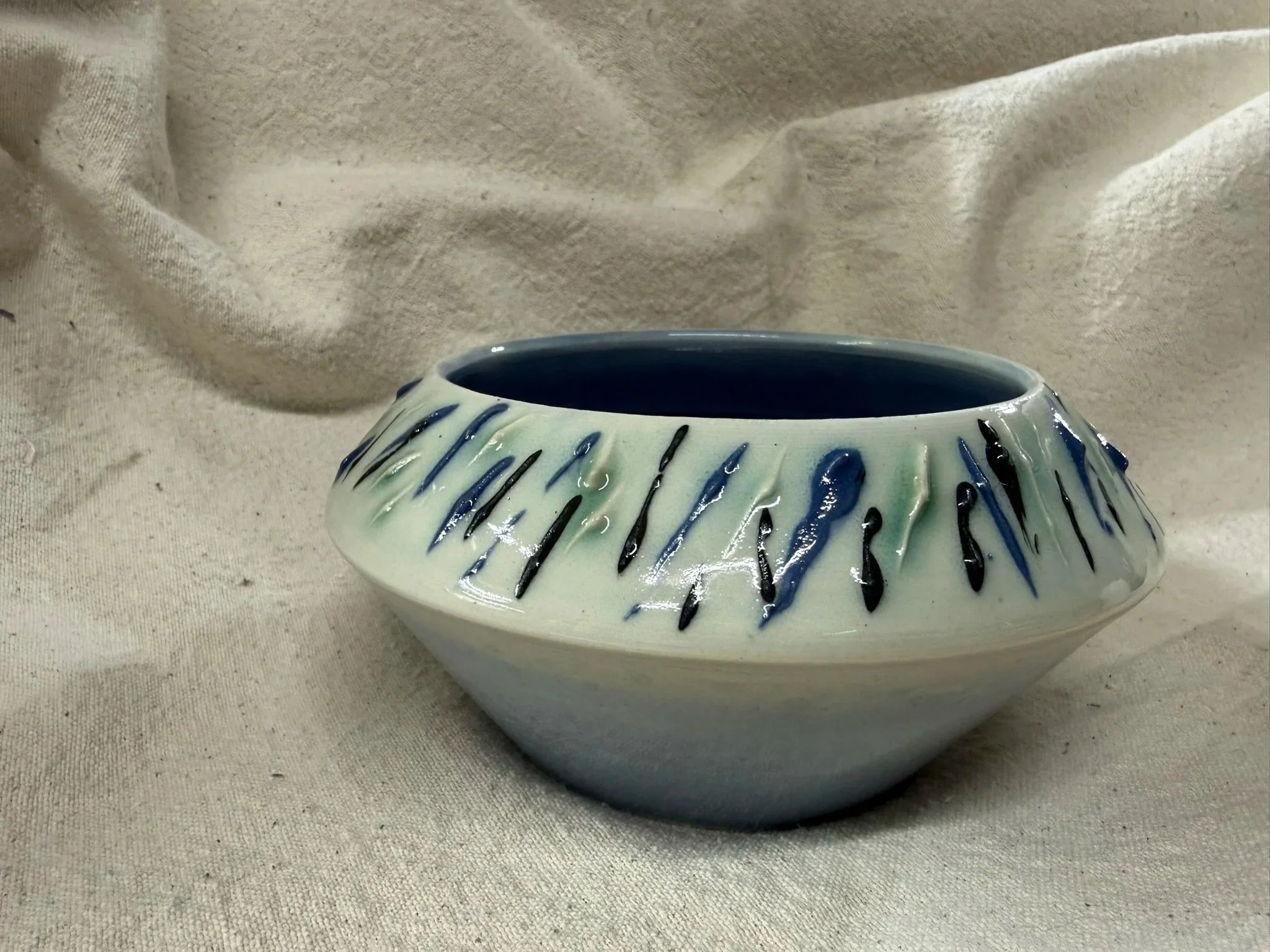 A ceramic bowl with a white glaze and blue and black drips decoration, placed on a light fabric surface.