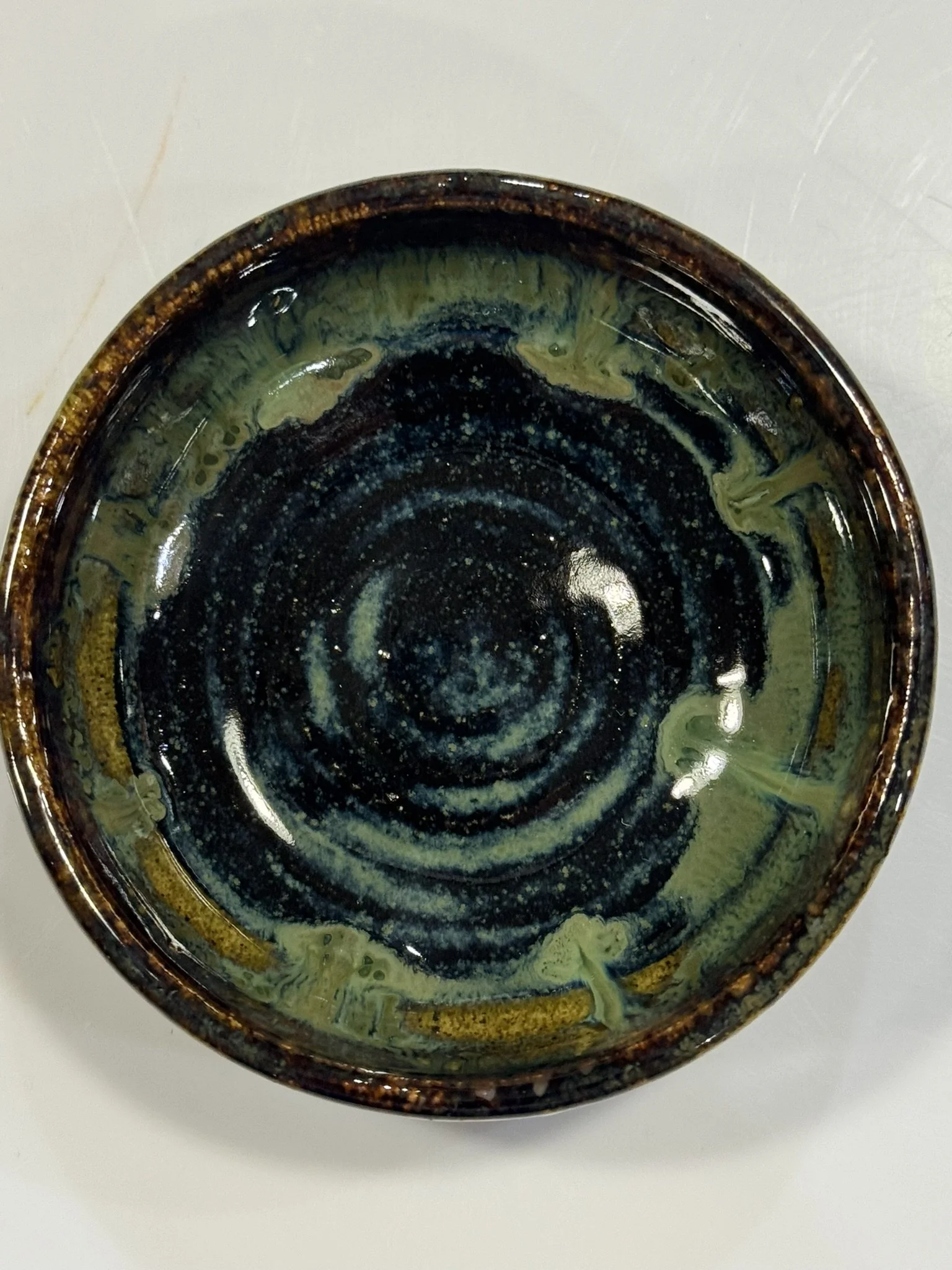 Top view of a ceramic bowl with a dark, glossy glaze and swirling yellow and green patterns inside.